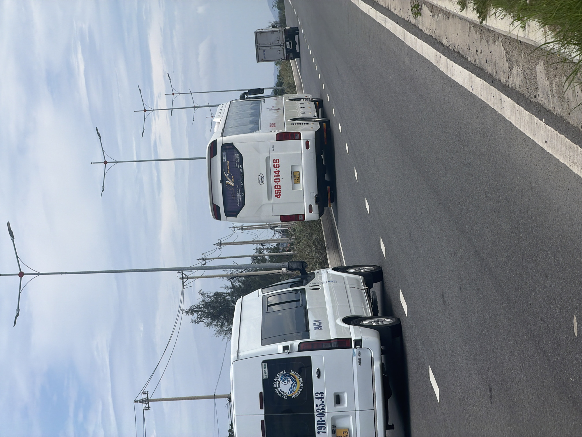 The media file captures a daytime scene on a multi-lane highway in Bac Cam Ranh Ward, Vietnam. The image is oriented vertically but depicts vehicles on a road. Two white passenger vehicles, resembling vans or small buses, are parked or driving on the rightmost lane, near the shoulder. A white truck with a cargo container is visible further down the road.
The sky is bright with scattered white clouds. To the left of the road, there are several tall light poles with multiple horizontal arms, as well as power poles carrying electrical lines. The road surface is asphalt with white dashed lane markings and a solid white line delineating the shoulder, which abuts a concrete curb and some sparse green vegetation. In the distant background, gentle mountains or hills are visible.
Visible text on the vehicles includes:
* On the white Hyundai bus: "XE DU LỊCH" (Tourist Car/Bus), "Vân Sơn Service", "IV-RSE" (part of the company name), and the license plate "49B-014.66".
* On the white passenger van: "XE HỢP ĐỒNG" (Contract Car/Bus), "OCEAN SMILE" logo, "CTY TNHH" (Limited Company), "HOTLINE- 079678979", "ABS" on the rear, and the license plate "79B-035.43".
* The truck in the distance has a partially visible license plate, likely "79L-582.53".