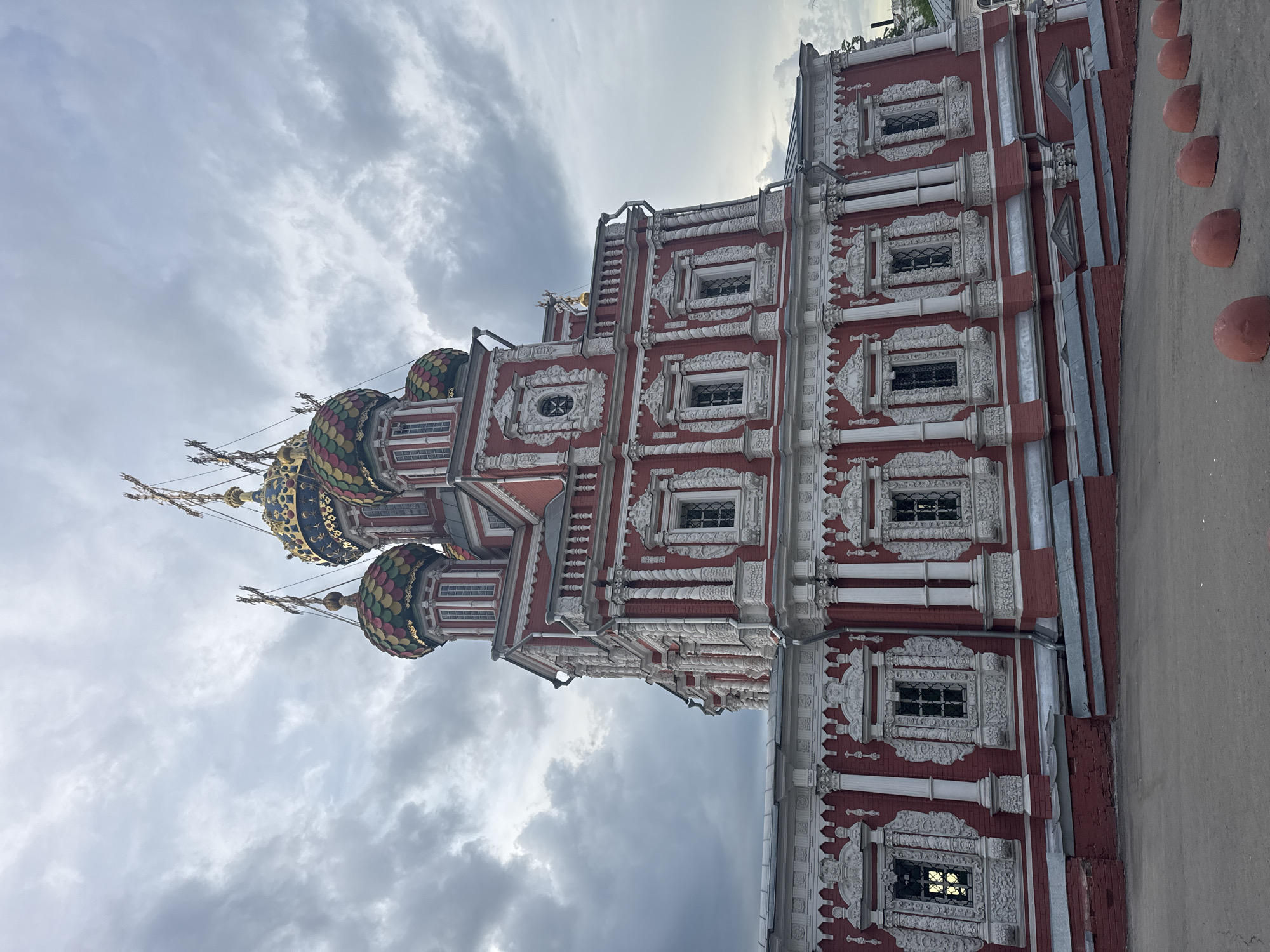 The image shows the Church of the Nativity of John the Precursor in Nizhny Novgorod, Russia. The church is a red and white brick building with ornate white detailing around the windows and edges. It has multiple domes, some of which are decorated with colorful scales and golden stars. The sky is cloudy and overcast. A line of rounded, terracotta-colored objects is visible along the bottom right edge of the image. The photograph is taken from a low angle, looking up at the church.