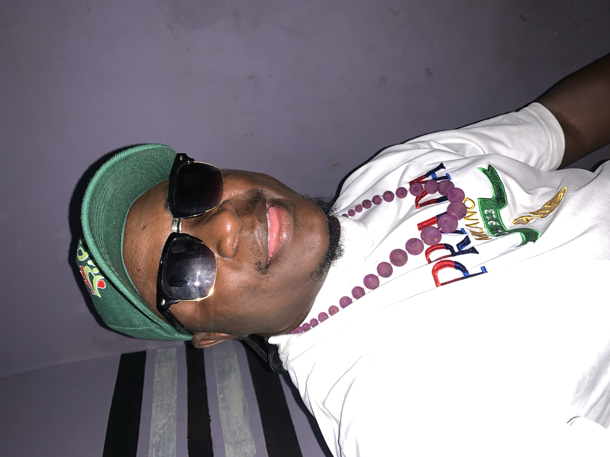 A person wearing sunglasses and a green hat is visible indoors. The person has dark skin, a slight beard, and is wearing a white T-shirt with colored embroidery and a purple beaded necklace. The background consists of a light purple wall with a striped pattern visible near the bottom of the frame. There is no visible evidence to support this image being taken in the city of Akwanga, Nigeria.