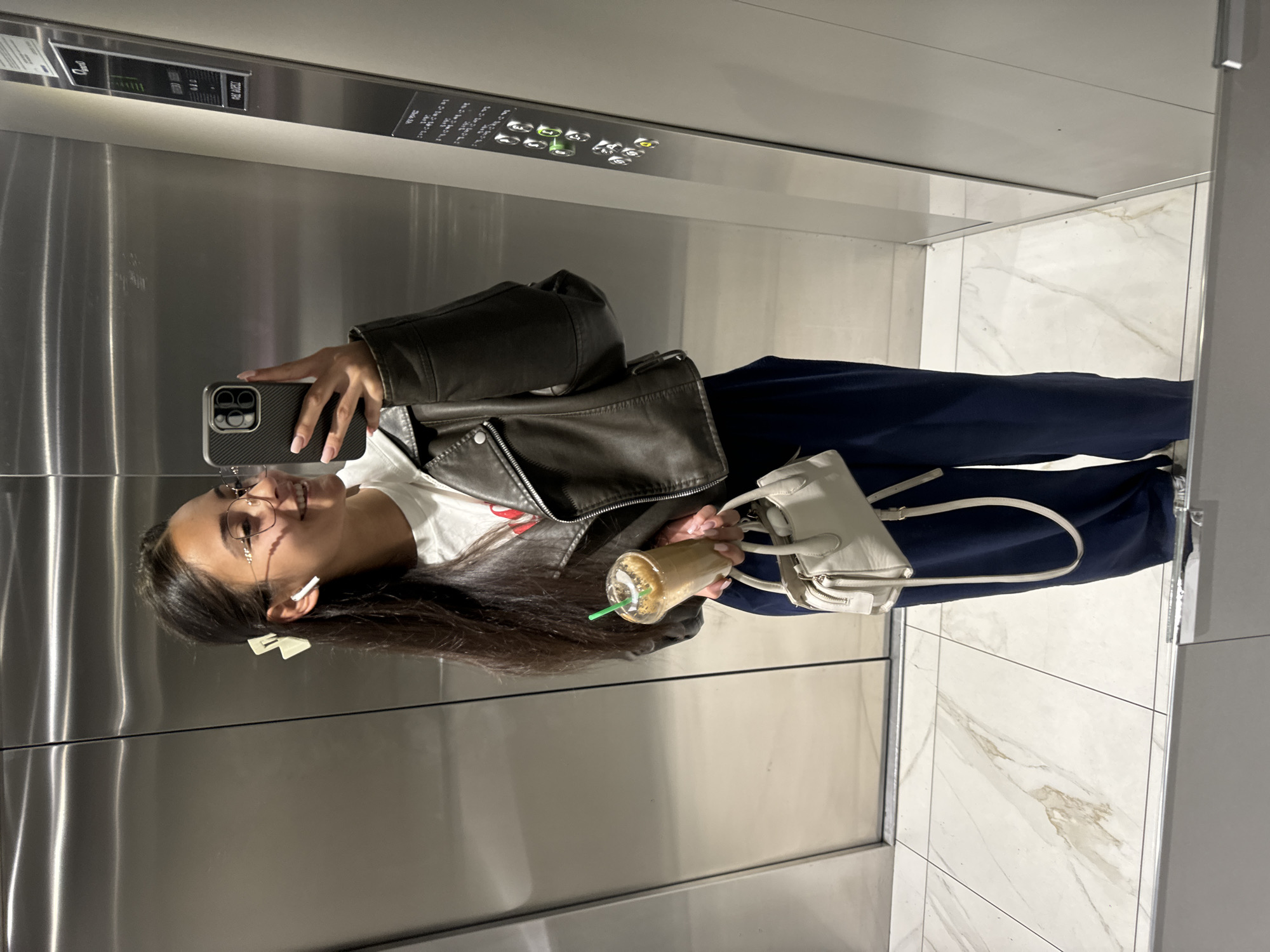 A woman is taking a selfie in an elevator. The elevator has a stainless steel interior with a control panel at the top. The floor of the elevator is tiled with white marble. The woman is wearing glasses, a white t-shirt, a black leather jacket, and navy blue pants. She is holding an iced coffee and a white purse. There is a white bow clip in her hair. The city of Sofia, Bulgaria, is not discernible from the image.