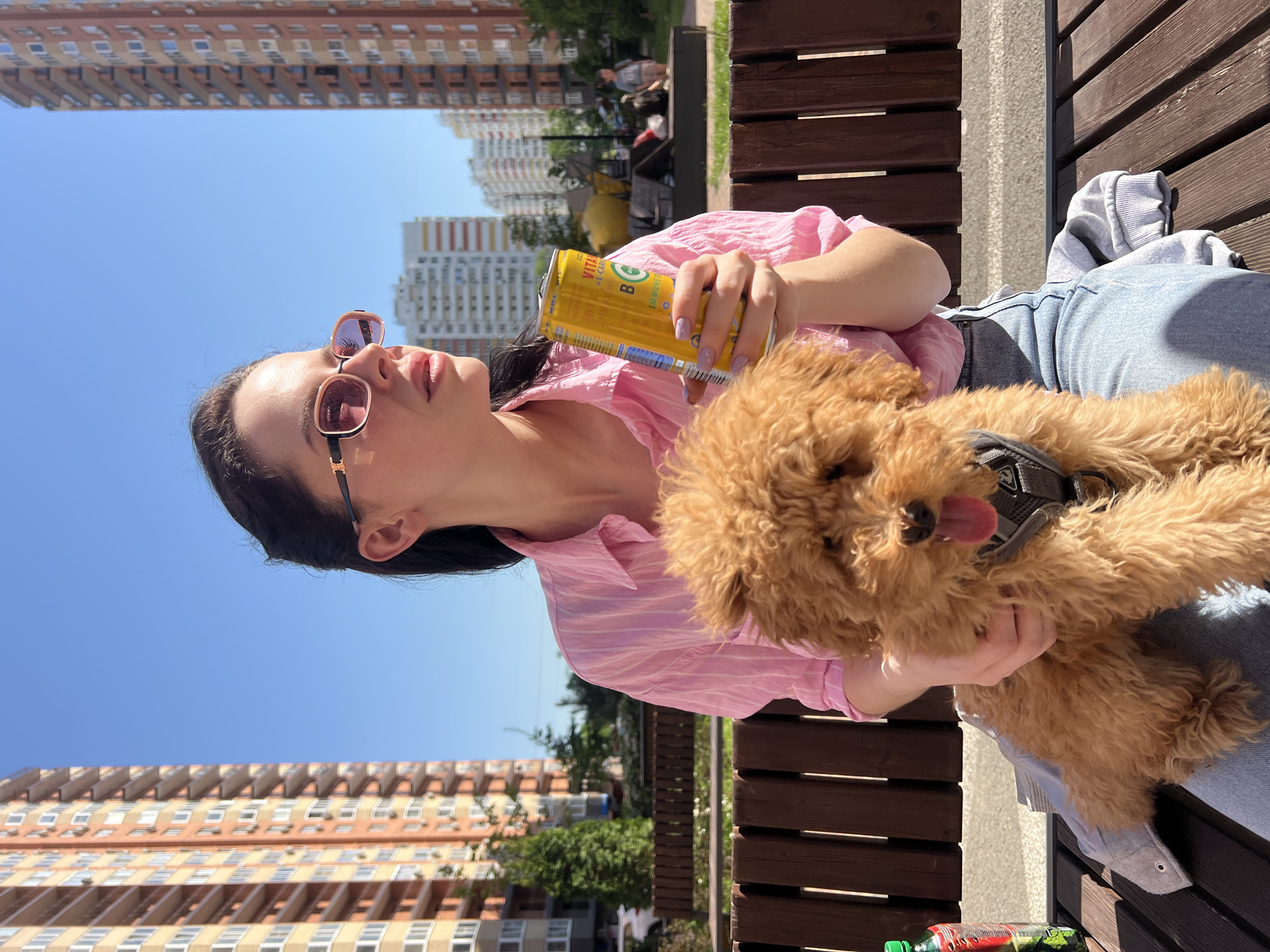 The image shows a woman sitting on a wooden bench in an outdoor setting. She is wearing pink sunglasses and a pink striped shirt, holding a yellow can of "Vita E-Carbo" beverage in one hand and a small, brown poodle in the other. The dog is wearing a harness and has its tongue sticking out. The woman appears to be looking upwards and slightly to the left.
Behind them, there are apartment buildings, trees, and a playground. The location is most likely in the city of Krasnodar, Russia, based on the design of the buildings and the brand of the beverage.