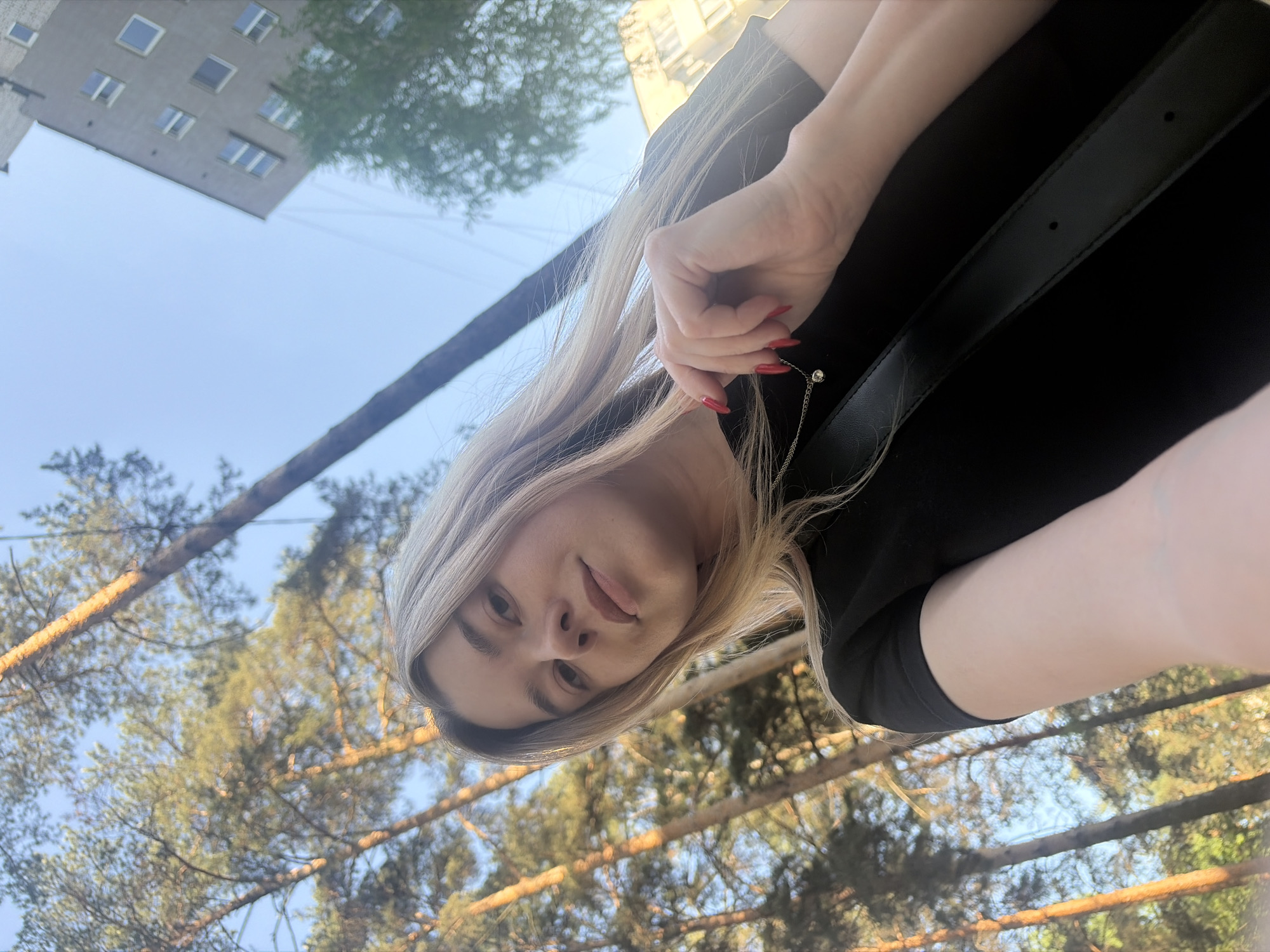 The image shows a woman with blonde hair, red nail polish, and wearing a black top with a strap, possibly from a bag. She appears to be taking a selfie from a low angle. In the background, there are trees and a tall building. Based on these details, the image could have been taken in a park or green space in the city of Chaikovsky, Russia.