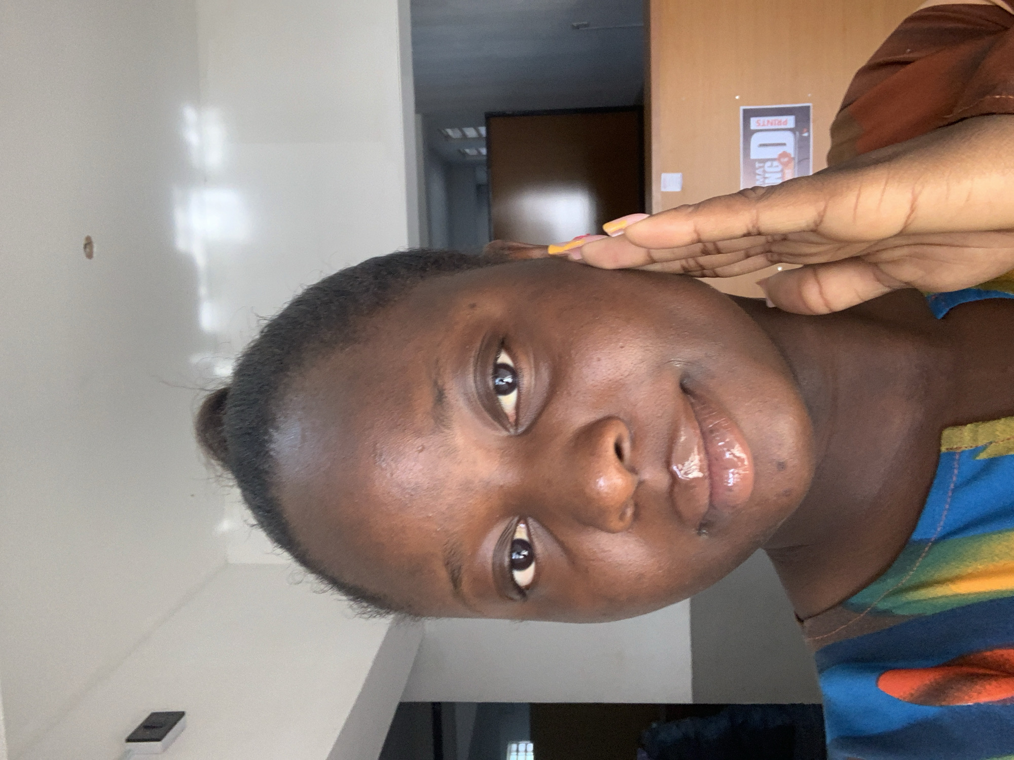 A close-up, medium shot captures a young Black woman from the chest up, posing with her right hand gently touching her face. Her dark hair is pulled into a bun on top of her head. She has brown eyes and full lips that appear to have lip gloss on them. Her skin is dark brown and smooth. She is wearing a colorful top with shades of blue, yellow, orange, and brown. The background is blurred and appears to be an indoor setting with a light-colored ceiling and a wooden counter. There is a black framed object mounted on the wall to the left and a sign on the wooden counter that reads "MAT NG D PRINTS". The lighting is bright and appears to be coming from above.