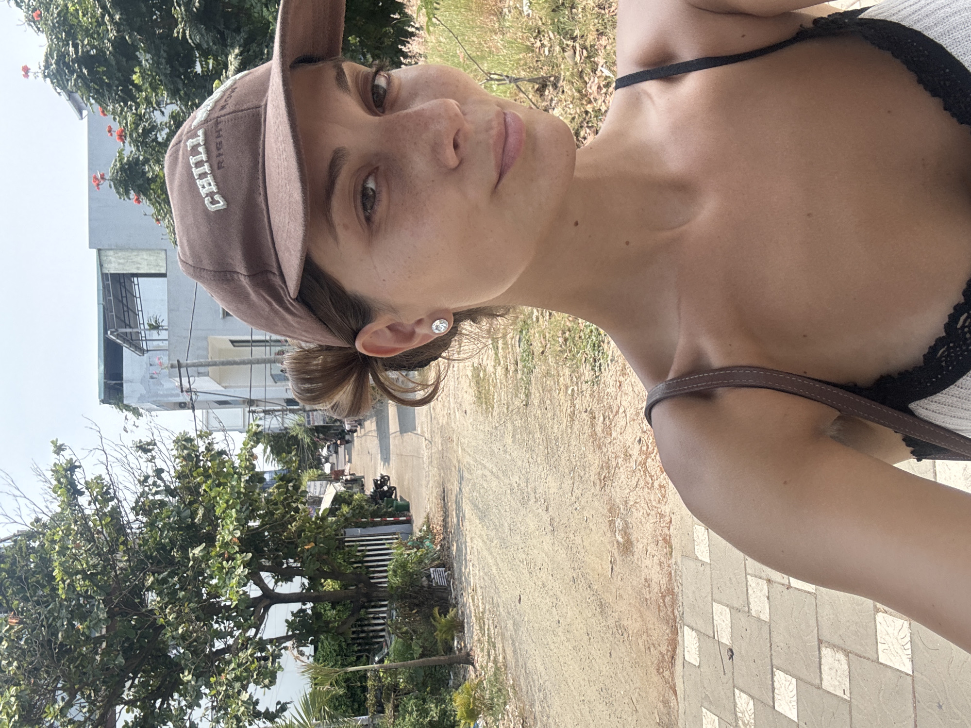 A woman in a brown baseball cap is standing on a paved walkway, taking a selfie. She is wearing a black spaghetti-strap top with a lacy trim and a brown shoulder strap. The backdrop reveals a dirt path with lush green trees and plants on one side and a building under construction on the other. The setting is likely within Đà Nẵng, Vietnam, given the tropical foliage and urban landscape.