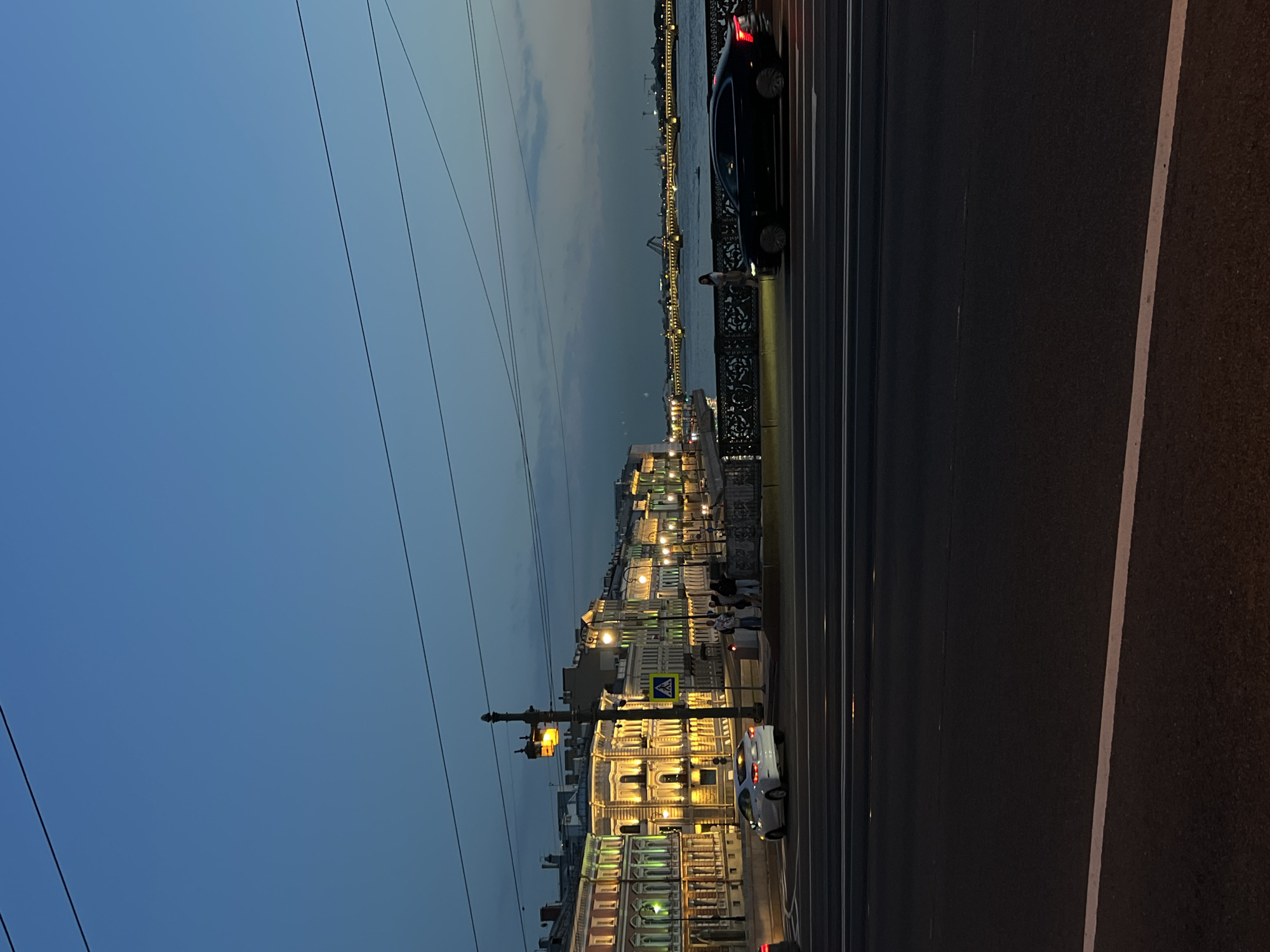 The image depicts a city scene at dusk, likely in Saint Petersburg, Russia, given the architecture and river. A wide asphalt road dominates the foreground with white lane markings. Beyond the road, a river flows, spanned by a bridge with decorative railings and illuminated by streetlights. On the far bank, ornate buildings with illuminated windows line the embankment, showcasing classical architecture with detailed facades. A few people are visible on the bridge and sidewalk. Several cars are present on the road, with one black sedan actively moving across the bridge and a white sedan stationary near an intersection. The sky is a deep blue transitioning to a lighter hue near the horizon, with subtle clouds. Overhead, power lines crisscross the sky. The lighting suggests it is evening, with artificial lights from the buildings and street lamps casting a warm glow.