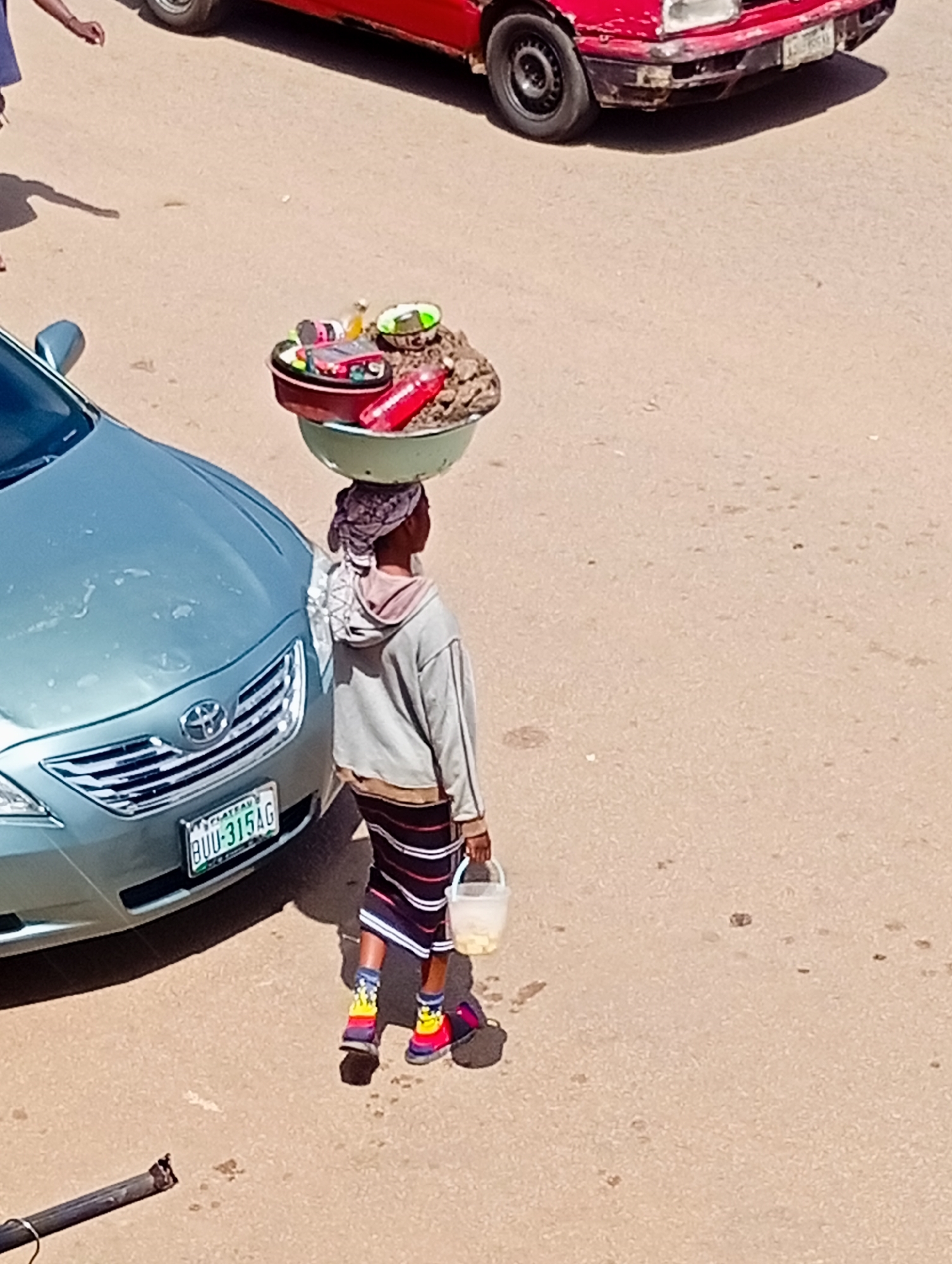 A person carrying goods on their head walks beside a light blue car on a paved street in Bukuru, Nigeria. The person, facing away from the camera, wears a grey hooded sweatshirt, a patterned skirt, colorful socks, and dark shoes. A white bucket is held in their right hand. The goods on their head are in a large, round, light green bowl. The light blue car has a license plate that reads "BUU-315AG". In the background, a red car is parked. The ground is paved and appears to be asphalt. The lighting suggests it is daytime.
