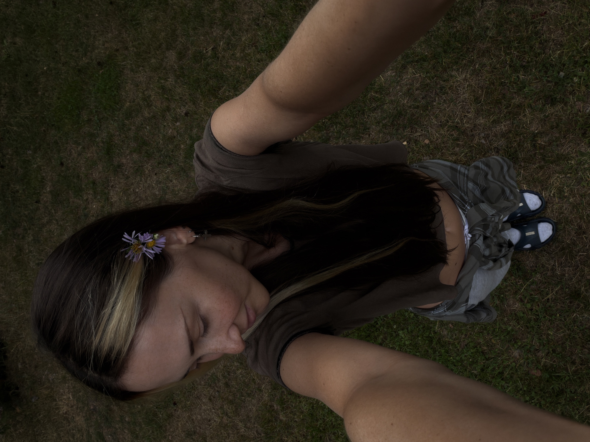 An overhead, high-angle, selfie-style, full-body shot shows a person with long dark hair, accented by a few blonde streaks and a small purple flower tucked behind their right ear. They are wearing a dark brown crop top, grey sweatpants with a green plaid shirt tied around their waist, white socks, and dark blue slides. The person is standing on a grassy area, likely a lawn or park, with dry patches and some green blades. The overall lighting is dim, suggesting it may be overcast or late in the day. The location is identified as Udelnaya, Russia.