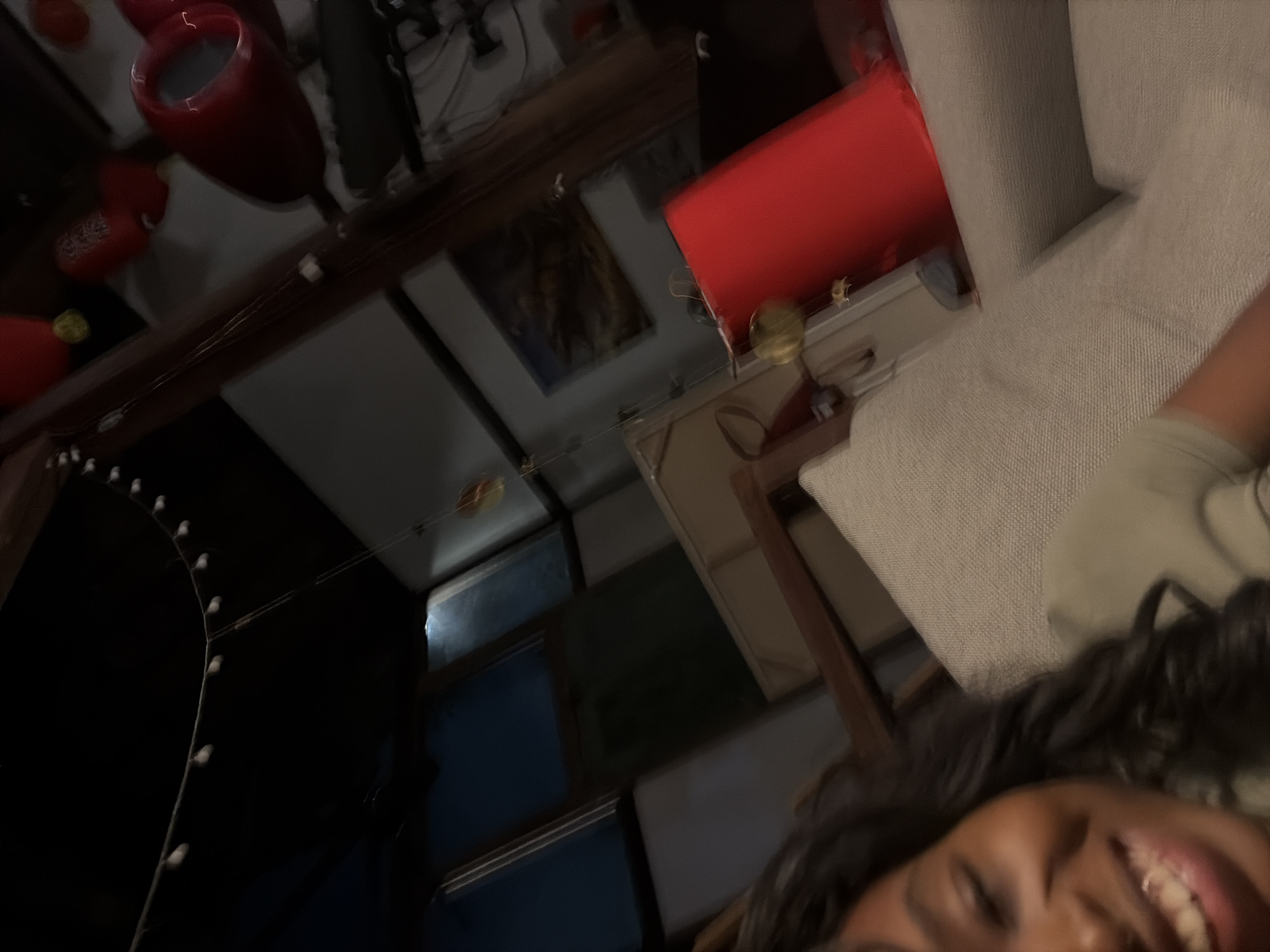 In this selfie-style photograph, a Black woman with dark curly hair is smiling broadly, her head and shoulders filling the left portion of the frame. She is seated on a textured light-colored sofa. In the background, the scene is dimly lit, suggesting an indoor setting. Several elements are visible, including a red cylindrical object on the right side of the image, possibly a lamp or a decorative piece. Above this, a framed artwork hangs on the wall, depicting what appears to be a landscape or abstract scene. Further into the background, there are windows with dark blue panes and possibly other decorative items, including some red lanterns hanging from the ceiling. The overall impression is one of a cozy, dimly lit interior space. The location in the city of Kediri, Indonesia, is not explicitly indicated by any visual cues within the image.
