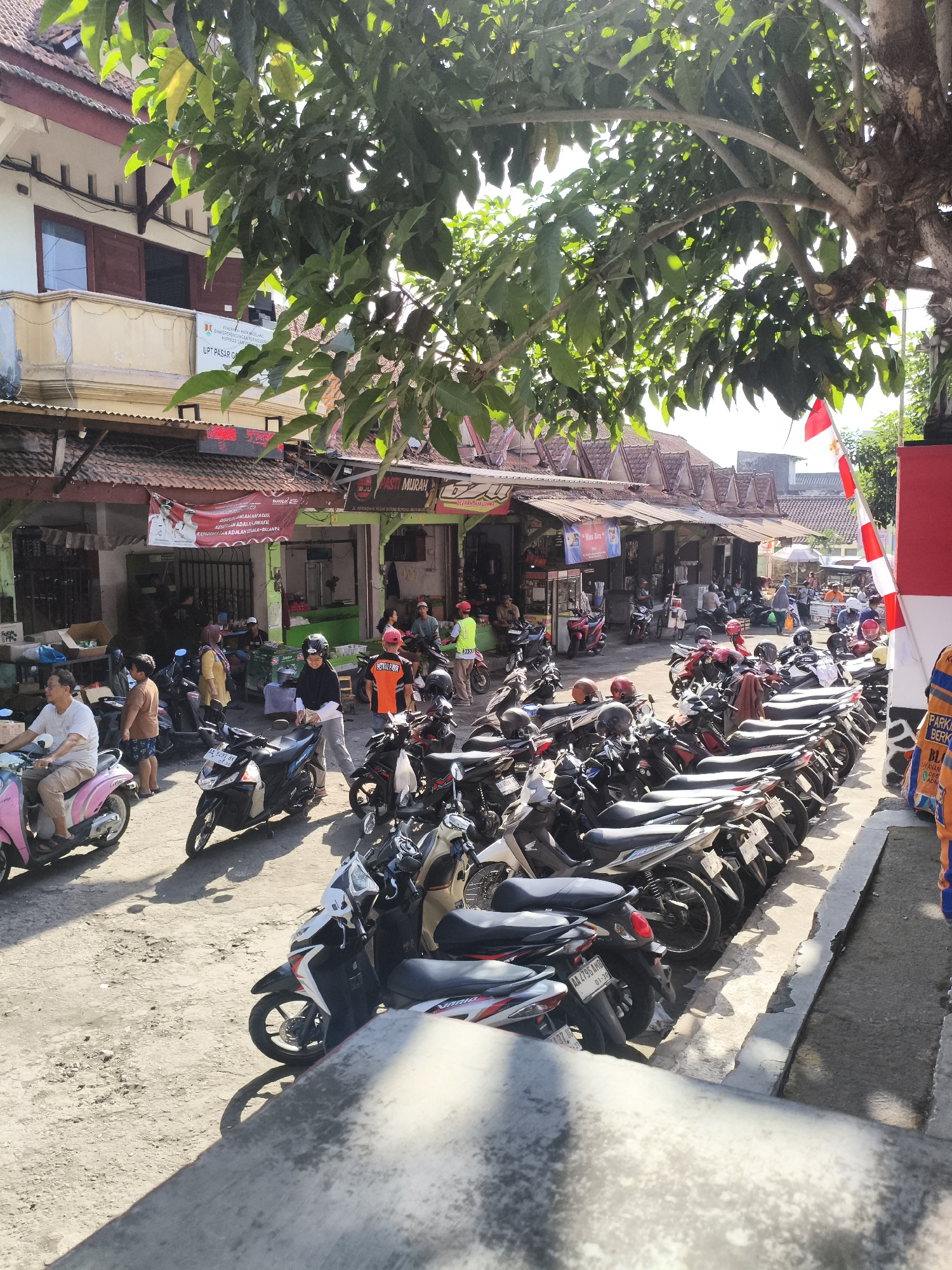 A sunny outdoor scene depicts a marketplace or street in Magelang, Indonesia, with a focus on numerous parked motorcycles and a row of shops. The ground is a dirt and gravel surface, illuminated by bright sunlight casting distinct shadows from the vehicles and overhead foliage.
In the foreground, a person in a light-colored shirt and dark pants is seated on a pink scooter. To their left, a person in a brown shirt and blue shorts stands.
A cluster of shops with corrugated metal roofs and open fronts lines the street. Banners and signs are visible, including one with red and white stripes and text in Indonesian. Some shops appear to be selling food or goods, with visible displays and people behind counters.
A significant number of motorcycles are parked in rows, filling much of the available space. These range from scooters to larger models, in various colors including black, white, red, and silver. Several people are interacting with the motorcycles; one person in a black helmet and black top is standing next to a black motorcycle, and another person in an orange vest is seen near a parked motorcycle.
Overhead, a large tree with green leaves provides shade, its branches extending across the upper part of the image. A red and white Indonesian flag is visible, attached to a pole on the right side of the frame. The architecture of the buildings suggests a traditional Indonesian market or commercial area. The overall impression is a busy, active marketplace during daylight hours.