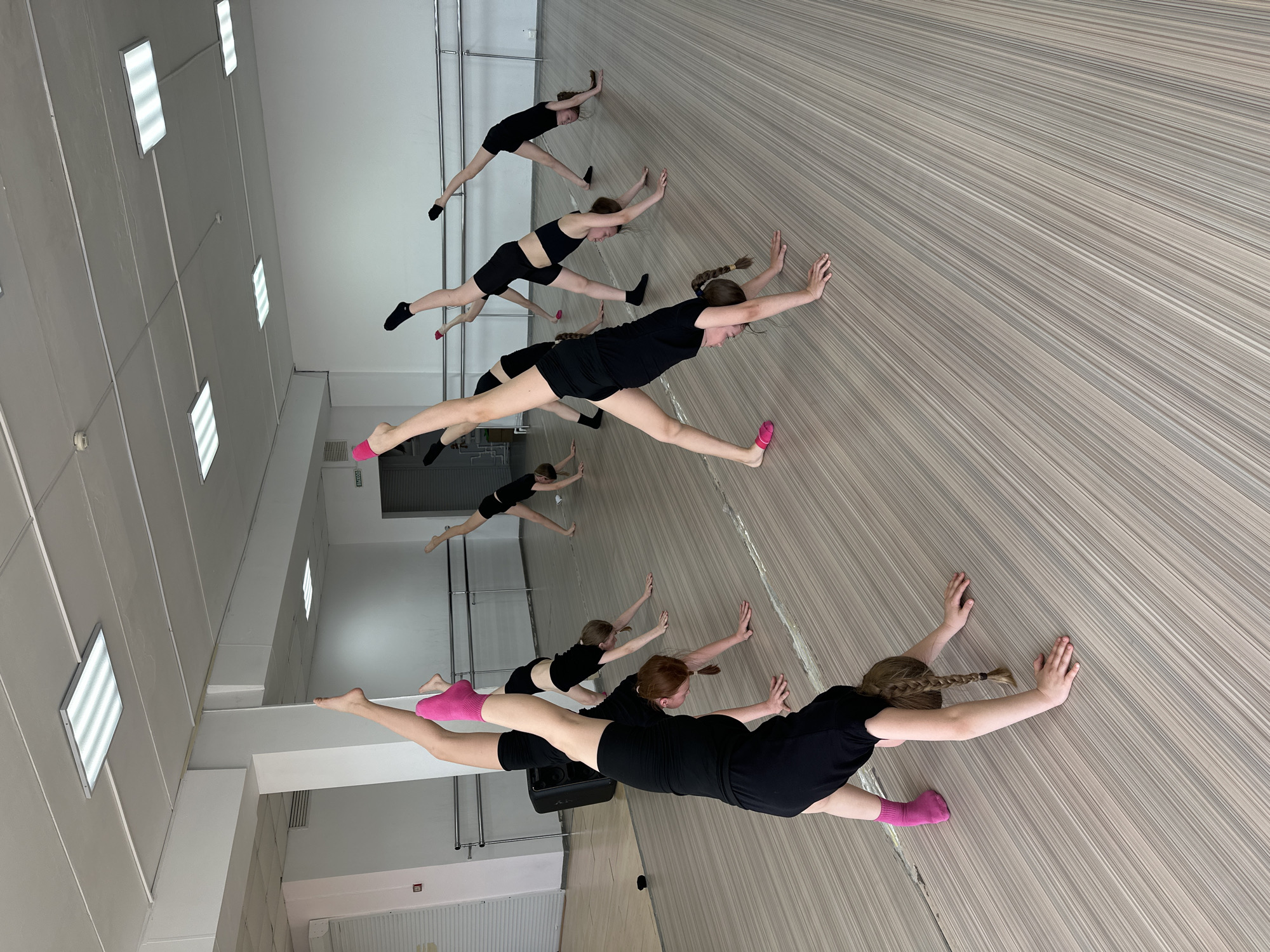 The image depicts a dance studio in Votkinsk, Russia, where several young women are practicing a stretching exercise. They are all clad in black dance attire, with some wearing pink or black socks. The studio has a light-colored wooden floor and white walls with square ceiling lights. The women are aligned, seemingly in a row, stretching their legs wide apart while supporting themselves with their hands on the floor.