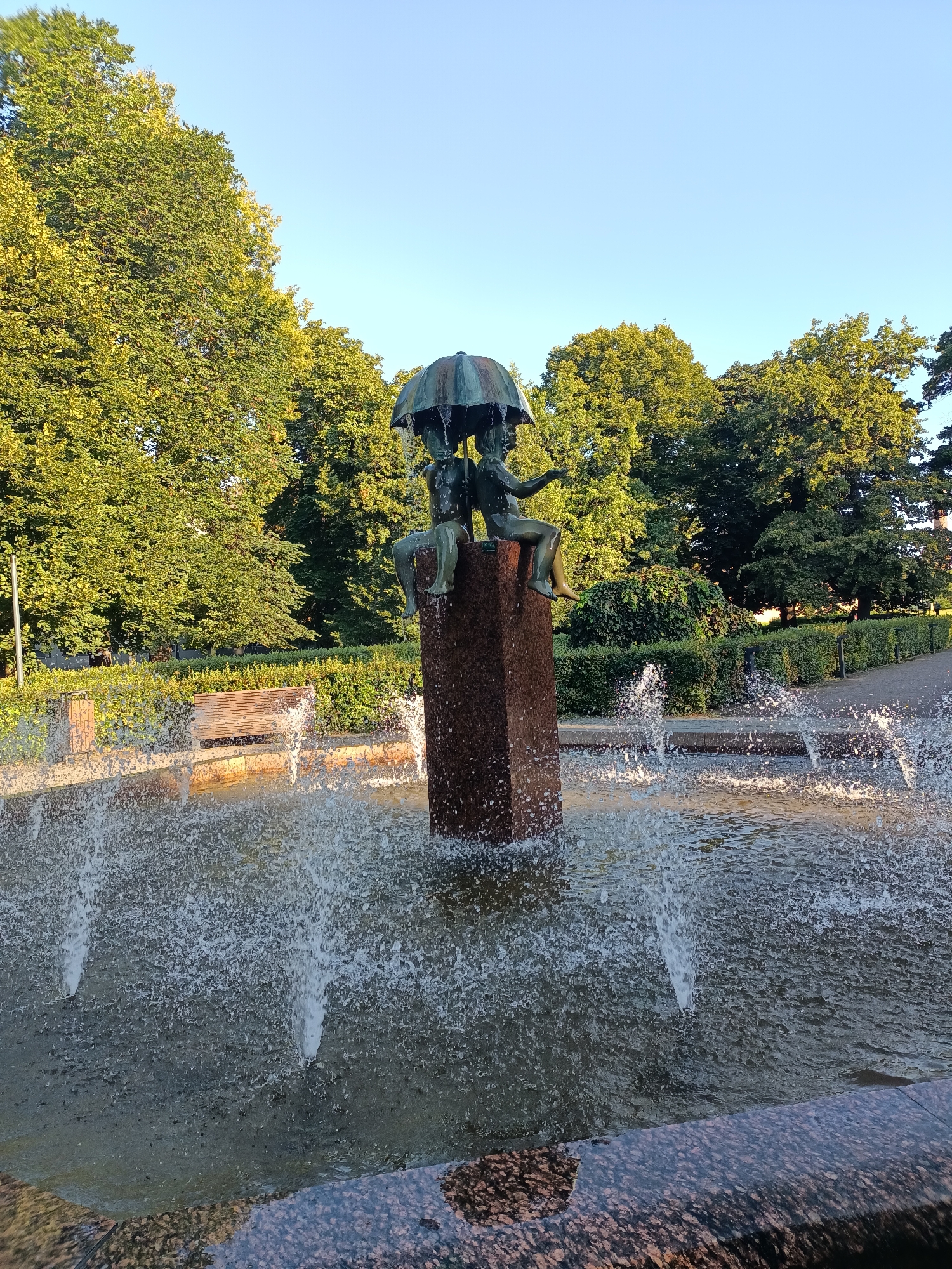 The image depicts a bronze sculpture of two children sharing an umbrella, situated atop a stone pedestal in a public fountain. The fountain is in motion, with water spraying upwards. The sculpture and fountain are surrounded by lush green trees and manicured hedges, suggesting a park or garden setting. In the background, a pathway and benches are visible. The sky is clear and blue, indicating a sunny day. This scene is located in the Kadriorg Park in Tallinn, Estonia.