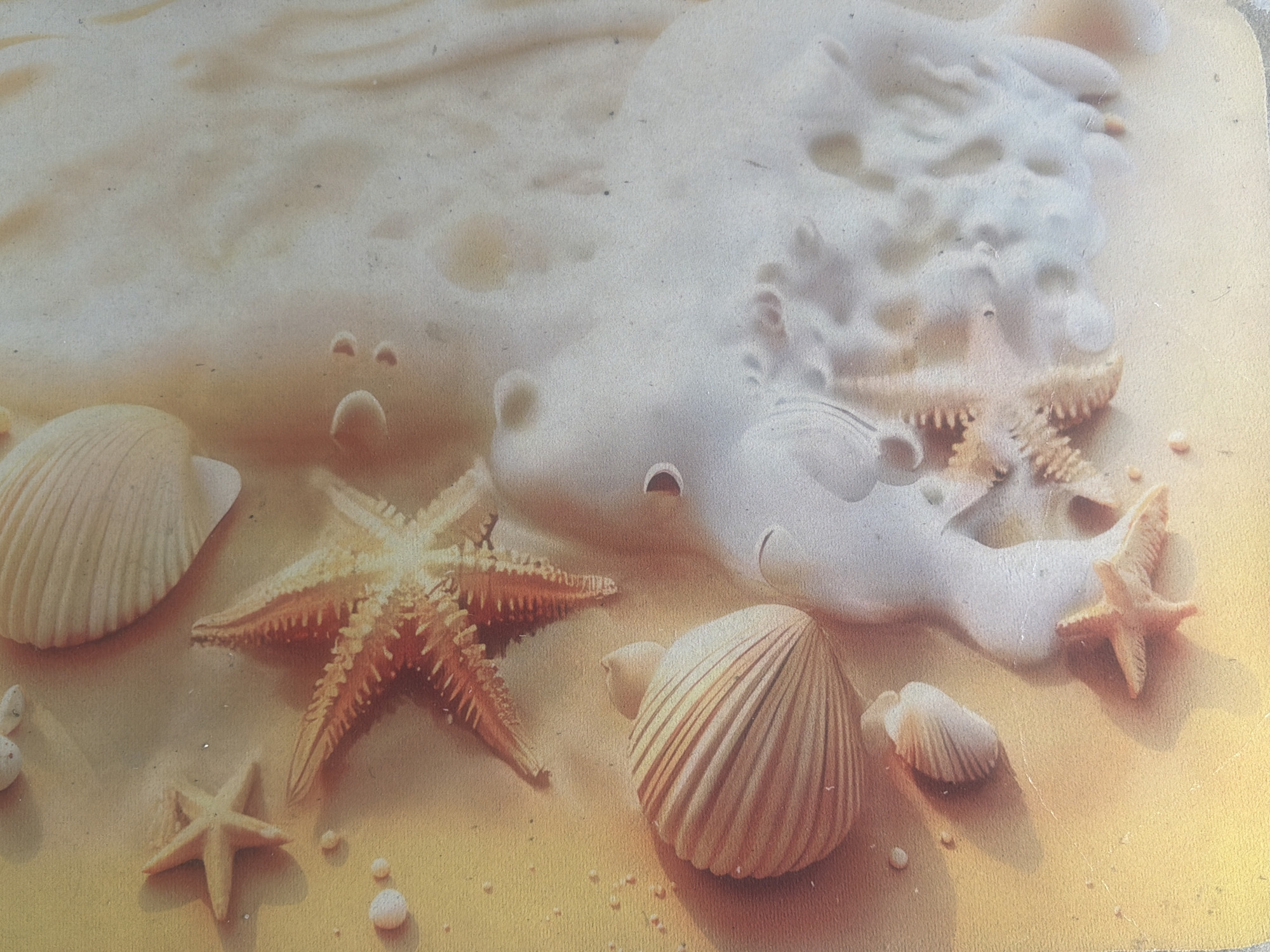 This is a close-up, high-angle photograph of a beach scene, likely a decorative representation rather than a real beach, given the glossy and somewhat artificial appearance. The subjects are seashells and starfish arranged on a sandy surface.
There are several distinct objects: a large, ridged seashell positioned on the left side of the frame, and a smaller, similar seashell towards the bottom right. Two prominent starfish are visible: a larger, darker-colored one in the center-left, and a smaller, lighter-colored one on the far right. There are also smaller, round white objects scattered across the sand, which could be pebbles or small shells.
The setting is a sandy beach, depicted with soft, warm tones of yellow and beige. A foamy wave, rendered in white, washes over part of the scene, creating a dynamic element. The wave's texture is soft and bubbly, suggesting a gentle surf.
There is no discernible activity taking place, as the scene is static, presenting a still-life arrangement of beach elements. The lighting appears to be soft and diffused, creating subtle shadows that add depth to the objects and sand. The overall mood is serene and evocative of a peaceful beach environment.
There is no visible text in the image. The scene's composition and the presence of artistic embellishments suggest it could be part of a placemat, a placque, or another decorative item. The mention of "Mandra, Greece" as a location context is not visually evident within the image itself.