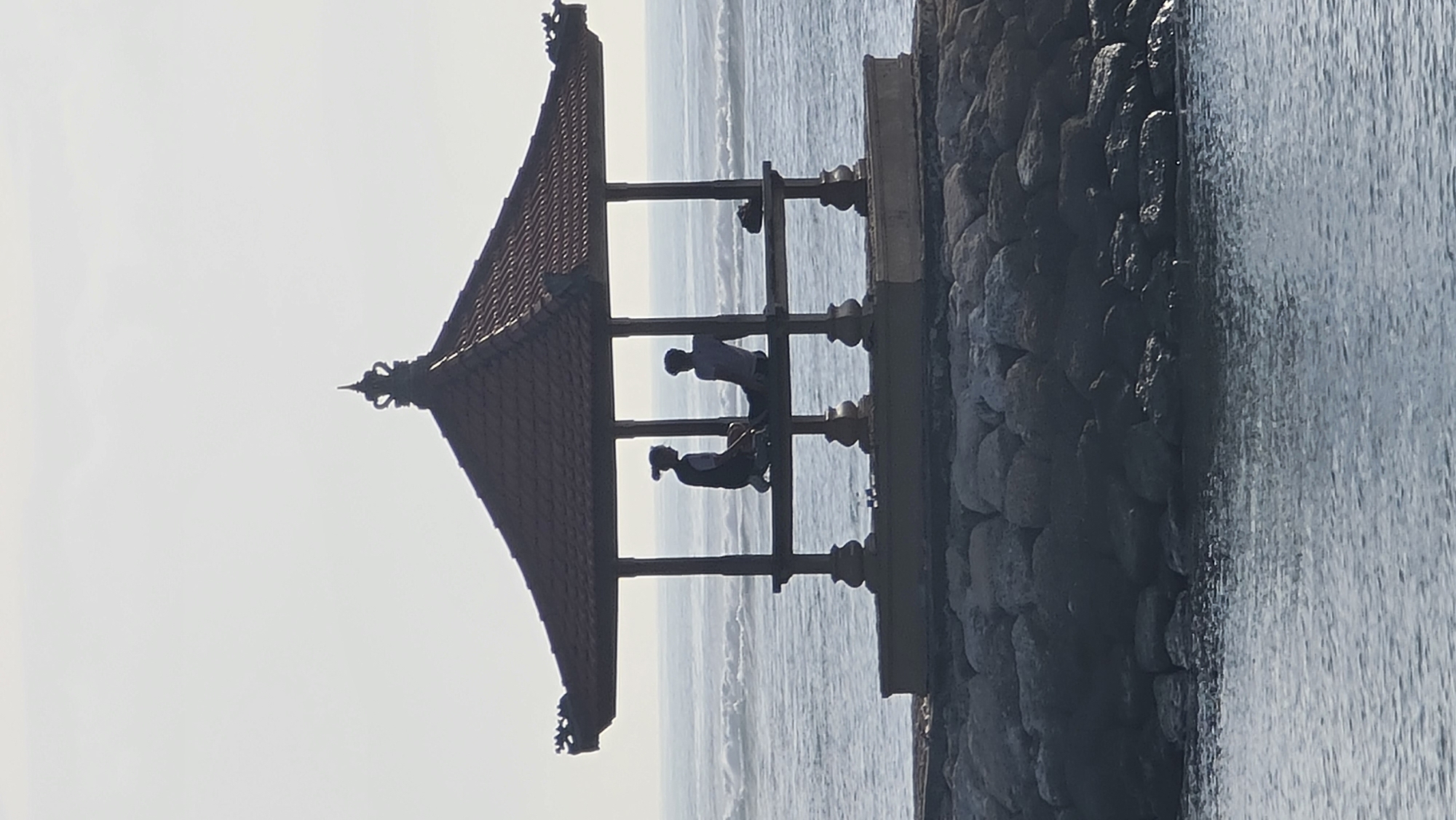 Two people are silhouetted inside a gazebo on a rocky embankment overlooking the ocean. The gazebo has a dark tiled roof with ornate trim and is supported by pillars. The person on the left is kneeling and facing the person on the right, who is seated. The ocean has moderate waves, and the sky is overcast. The gazebo appears to be a traditional Balinese structure, suggesting the location is a coastal area in Denpasar, Indonesia.