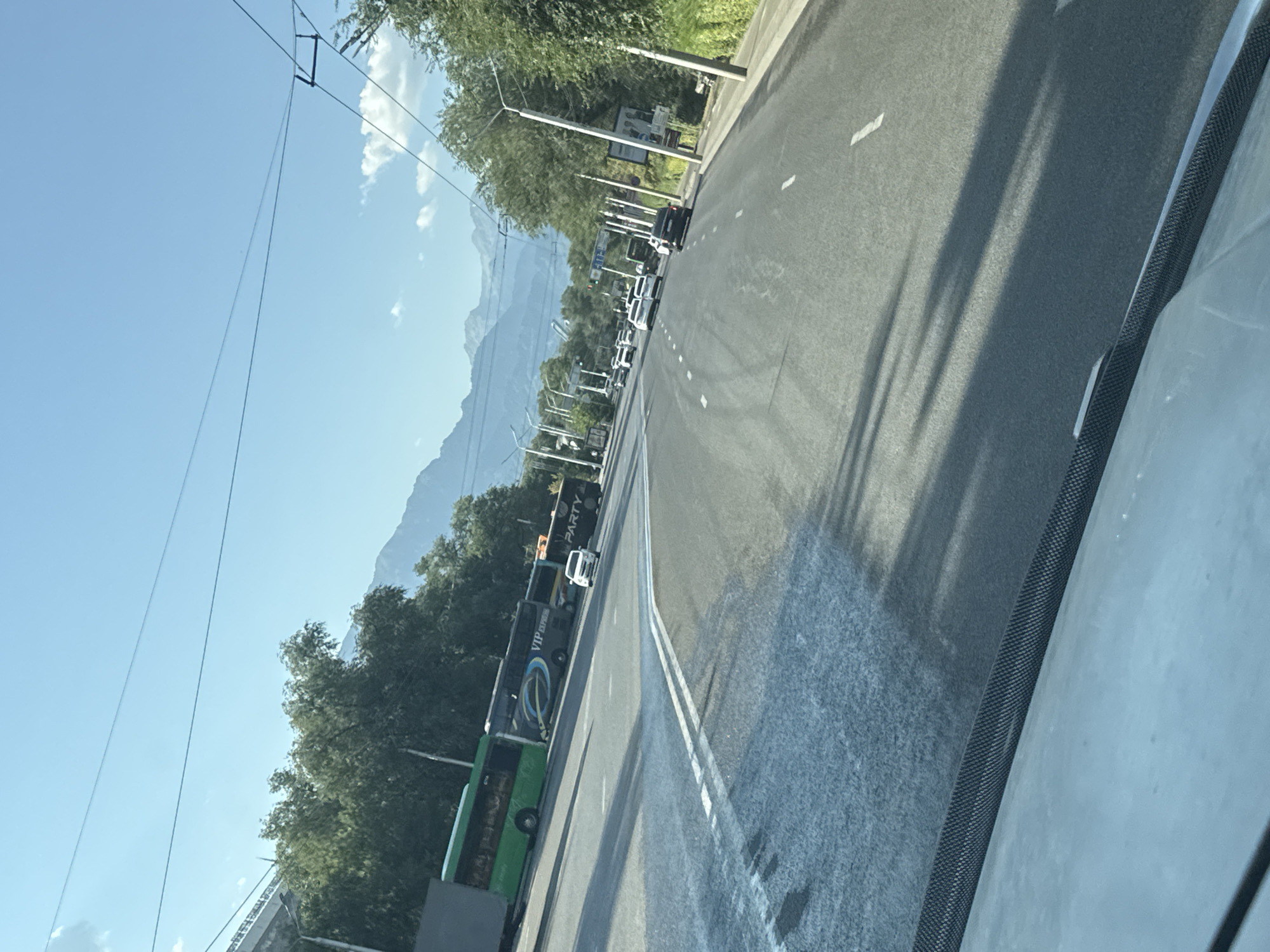 The image depicts a street scene in Almaty, Kazakhstan, with mountains visible in the background. Multiple buses are parked on the left side of the road, including a green bus and a dark bus with "VIP EXPRESS" branding. Several cars are driving on the multi-lane asphalt road. The sky is clear and blue. The perspective suggests the photo was taken from inside a vehicle moving along the road. The presence of overhead wires indicates an urban environment with possibly public transportation like trams or trolleybuses. The time of day appears to be daytime with ample sunlight casting shadows.