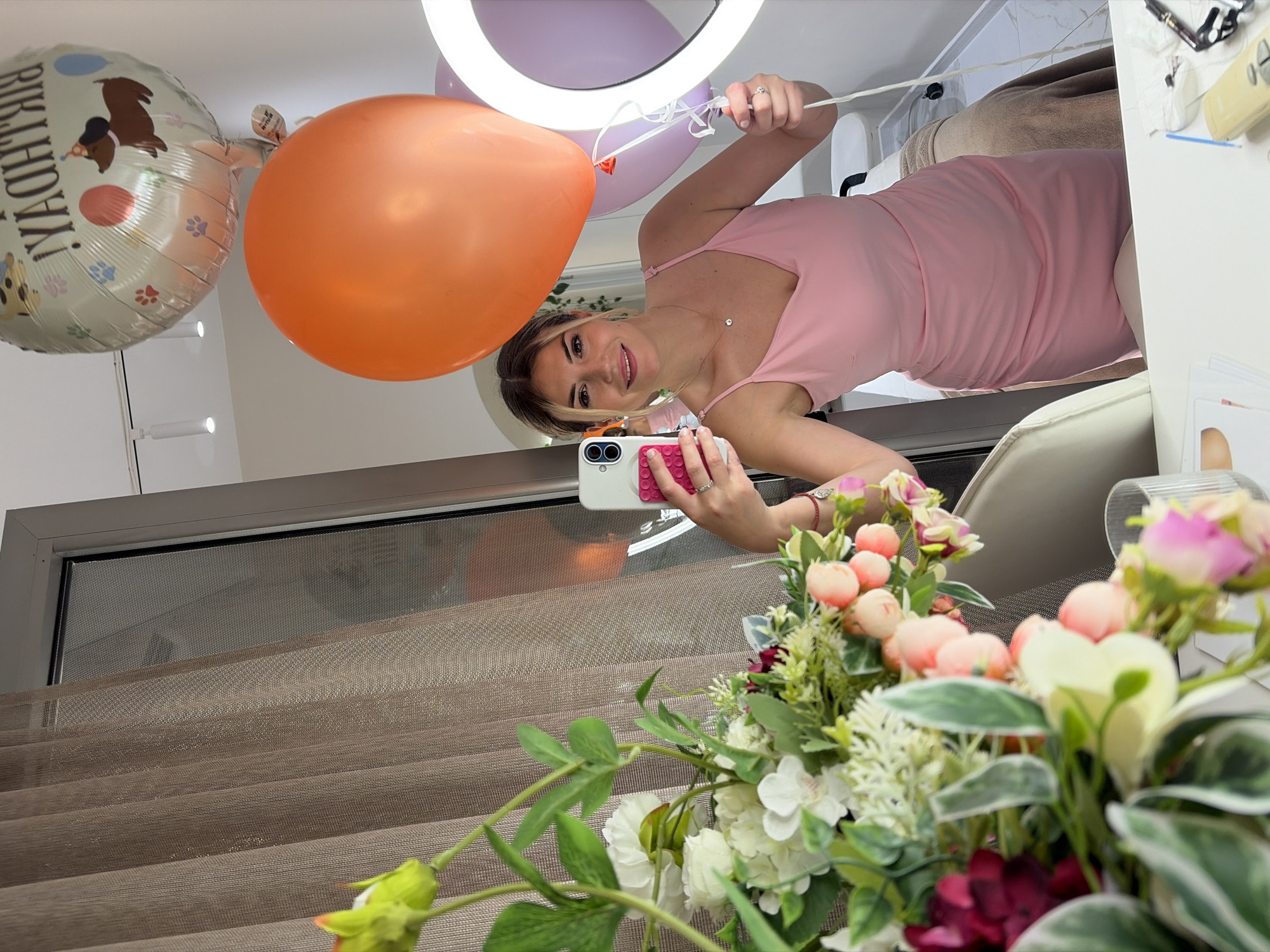 A woman, likely in her late 20s or early 30s, is taking a selfie in what appears to be a salon or beauty studio. She is wearing a pink, form-fitting dress with spaghetti straps. Her blonde hair is styled in loose waves, and she is smiling at the camera. She is holding a smartphone in her right hand, with a pink popsocket attached. In her left hand, she is holding the strings of several balloons: a large orange balloon, a lilac balloon, and a foil balloon decorated with a cartoon dog and the words "Happy Birthday" (inverted in the reflection).
The setting includes a large mirror, reflecting the woman and the balloons. To the left of the woman in the reflection is a curtain, and to the right, a ring light is visible, suggesting a professional photography or videography setup. In the foreground, a large bouquet of artificial flowers, primarily white and pink roses with green foliage, is positioned to the left. On a table in the foreground, partially visible, are various beauty products and tools, including a small white remote control, tweezers, and what appears to be a roll of gauze.
The overall ambiance is festive due to the balloons, suggesting a birthday celebration. The location is not explicitly Budva, Montenegro, but the context suggests an indoor, possibly commercial establishment.