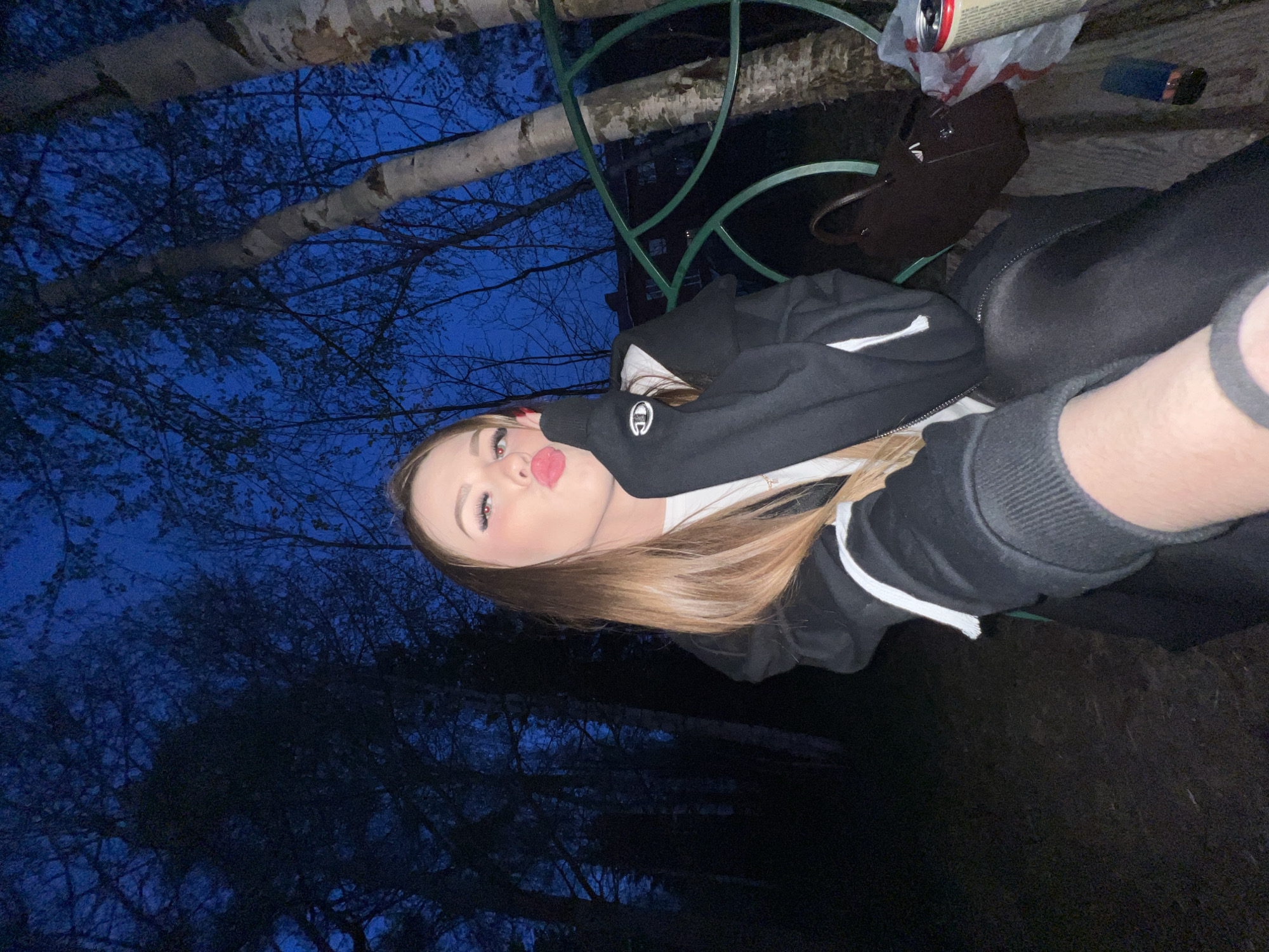 A selfie captures a young woman with long blonde hair, pursing her lips, in an outdoor setting at dusk or night. She is wearing a black hoodie with white stripes and a white shirt underneath. In the background, there are trees with bare branches against a dark blue sky. A metal railing is partially visible, as well as a few items including a can and a brown purse. The location is Noviy, Russia.