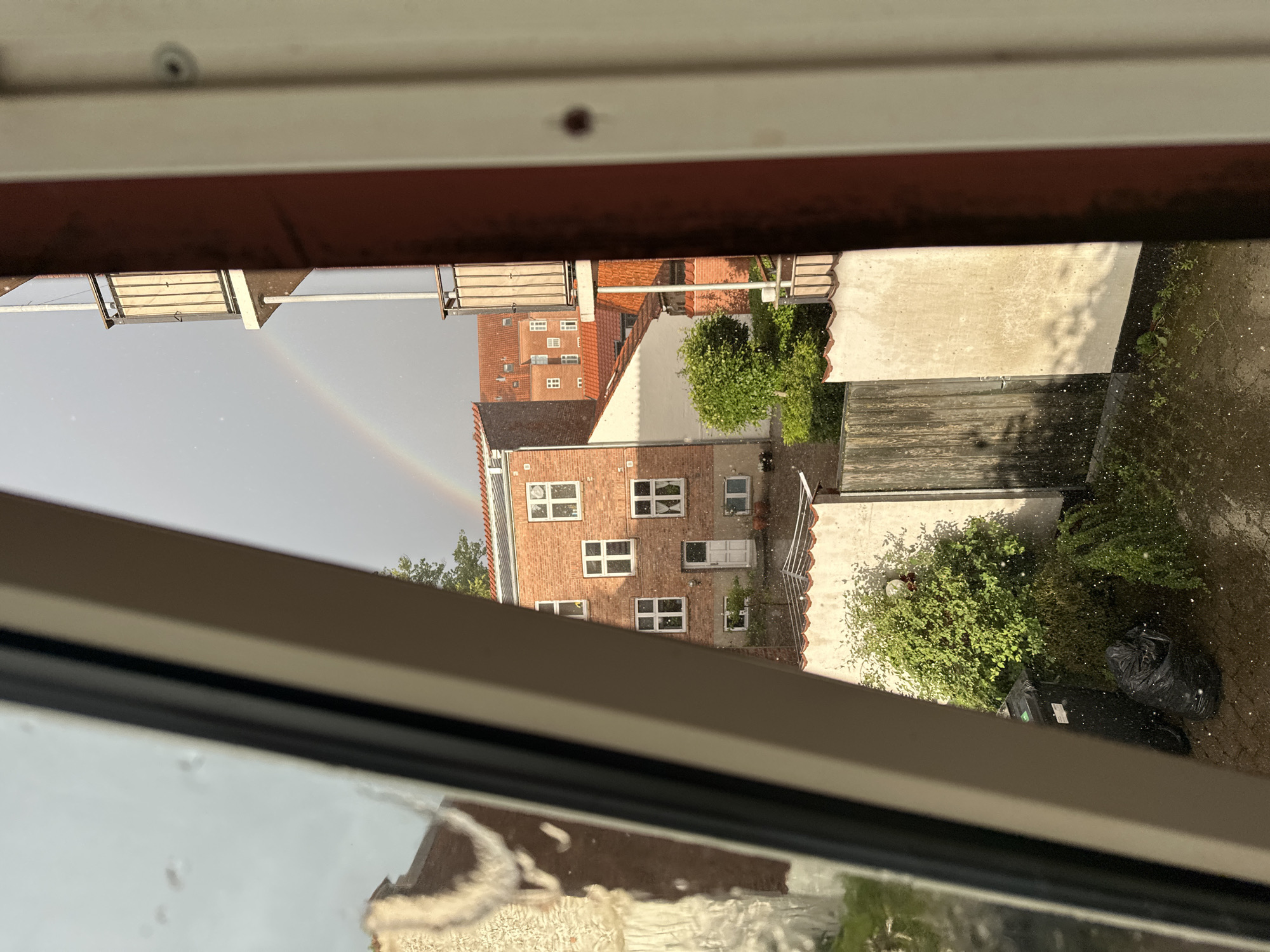 The image depicts a view through a window in Horsens, Denmark, on a rainy day with a rainbow visible in the sky. In the foreground, the edge of a window frame and glass is visible, with raindrops on the exterior. Beyond the window, a red brick building with white-framed windows and a red tiled roof is prominent. A balcony extends from an adjacent building on the right. In the lower part of the scene, a black trash bin and a black garbage bag are on a paved area. Green bushes and trees are present, and water is pooling on the ground. The overall scene captures a residential area in Horsens during a rain shower and the appearance of a rainbow.
