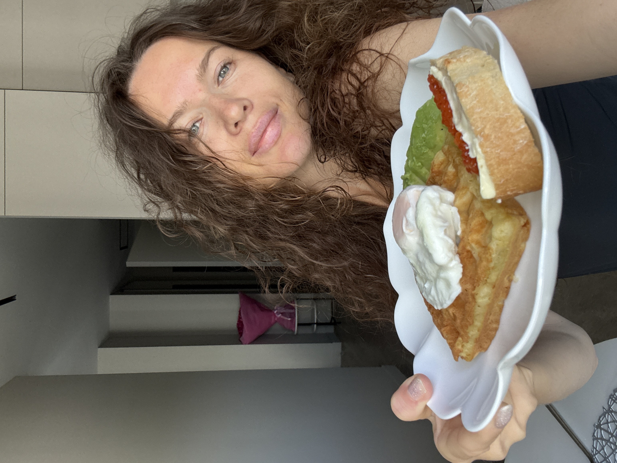 A woman with long, curly brown hair is holding a white, flower-shaped plate with breakfast in front of her face. The breakfast consists of a waffle topped with a poached egg, mashed avocado, and a slice of baguette with butter and red caviar. The woman is looking towards the camera with a slight smile. The background appears to be an indoor setting, possibly an apartment in Khimki, Russia. A bouquet of pink flowers is visible on a stand in the background, along with architectural elements of a modern interior. The lighting suggests it is daytime.