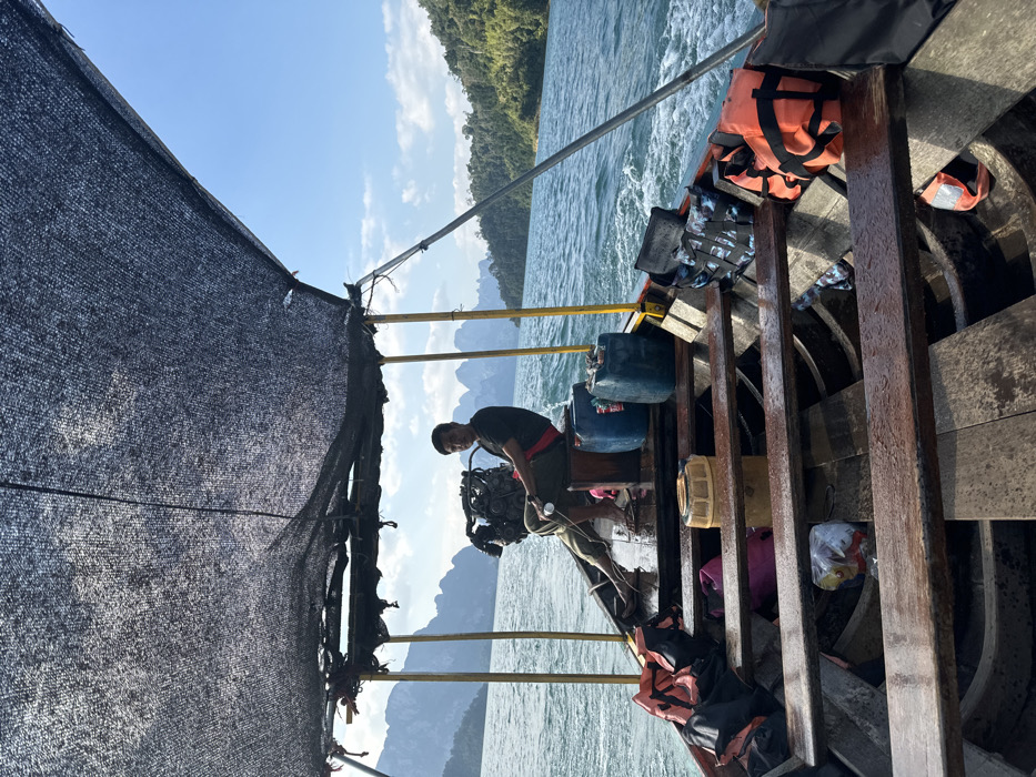 A longtail boat travels across a body of water under a clear blue sky. The scene is observed from inside the boat, looking towards its stern. A male operator, wearing a black shirt, dark pants, and a red sash, is seated barefoot on a wooden platform at the stern, operating a visible longtail engine with his right hand. Two blue plastic jerry cans are positioned near him. The boat's interior features multiple wooden crossbeams serving as benches and a wet wooden floor. Various items are stowed within the boat, including orange and camouflage-patterned life jackets, a pink bag, a white plastic bag, and a lidded cylindrical container. A dark mesh canopy provides shade overhead, supported by a basic framework. In the background, numerous steep, vegetated limestone karst formations rise from the water, characteristic of a tropical lake or reservoir environment. The water surface is choppy, with a visible wake trailing the boat.