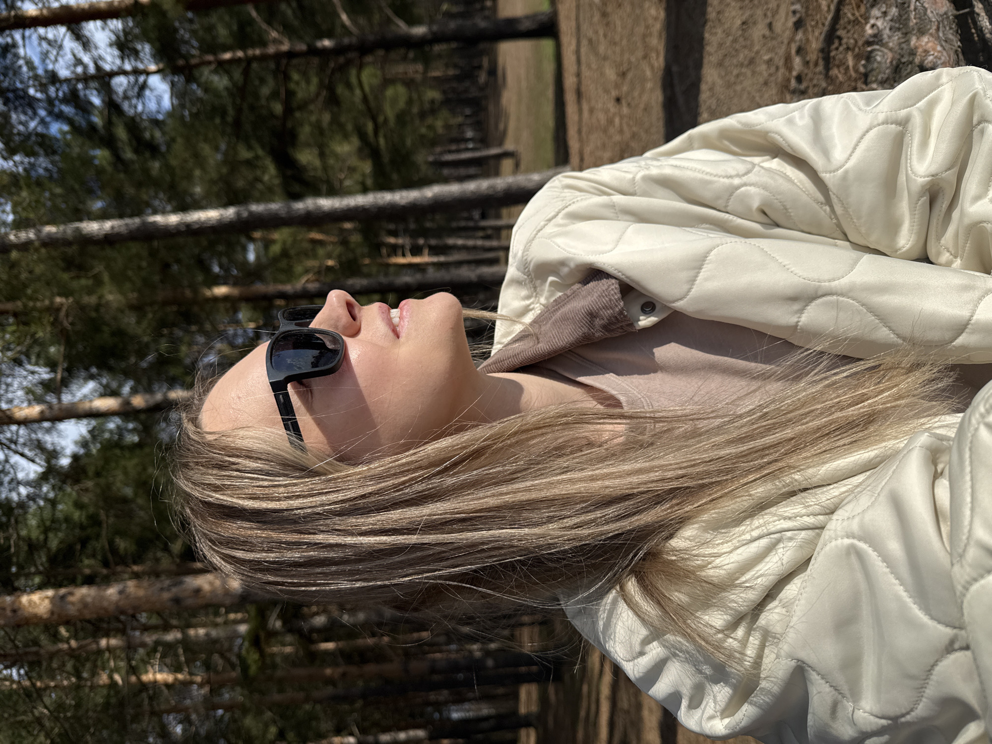 The image captures a close-up profile of a young woman situated in a sunlit coniferous forest. She is looking upwards, with her mouth slightly agape and a relaxed expression. Black sunglasses are perched on her nose. Her long, straight, blonde hair cascades over her shoulder and back, blending with her light cream-colored, quilted jacket. A brown or beige top is visible beneath the jacket.
The background features numerous tall, slender tree trunks, characteristic of a pine forest, extending upwards. Several horizontal logs or fallen trees are visible in the middle distance, suggesting a natural and undisturbed woodland environment. The ground appears to be covered with dry earth and pine needles.
The scene is bathed in bright, natural daylight, indicating a sunny day, likely late morning or afternoon, given the strong illumination and visible shadows. The clear weather contributes to a serene and pleasant atmosphere. The woman appears to be engaged in a moment of leisure, possibly enjoying the warmth of the sun or contemplating the canopy above. The setting is consistent with a forest in Chaikovsky, Russia. No visible text is present in the media.