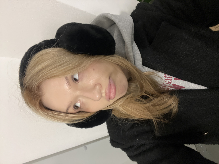A close-up, chest-up portrait depicts a young individual with light blonde hair, wearing black fuzzy earmuffs, a light grey hooded sweatshirt, and a textured black coat. The individual's head is tilted slightly, and their light-colored eyes are directed upwards and to the left. Their lips are pursed in a pout. A white graphic with red text is partially visible on the grey sweatshirt below the chin. The background consists of a plain, light-colored wall with a subtle corner or edge visible on the right and a small, dark circular detail on the left. The scene is located in the city of Chaikovsky, Russia.