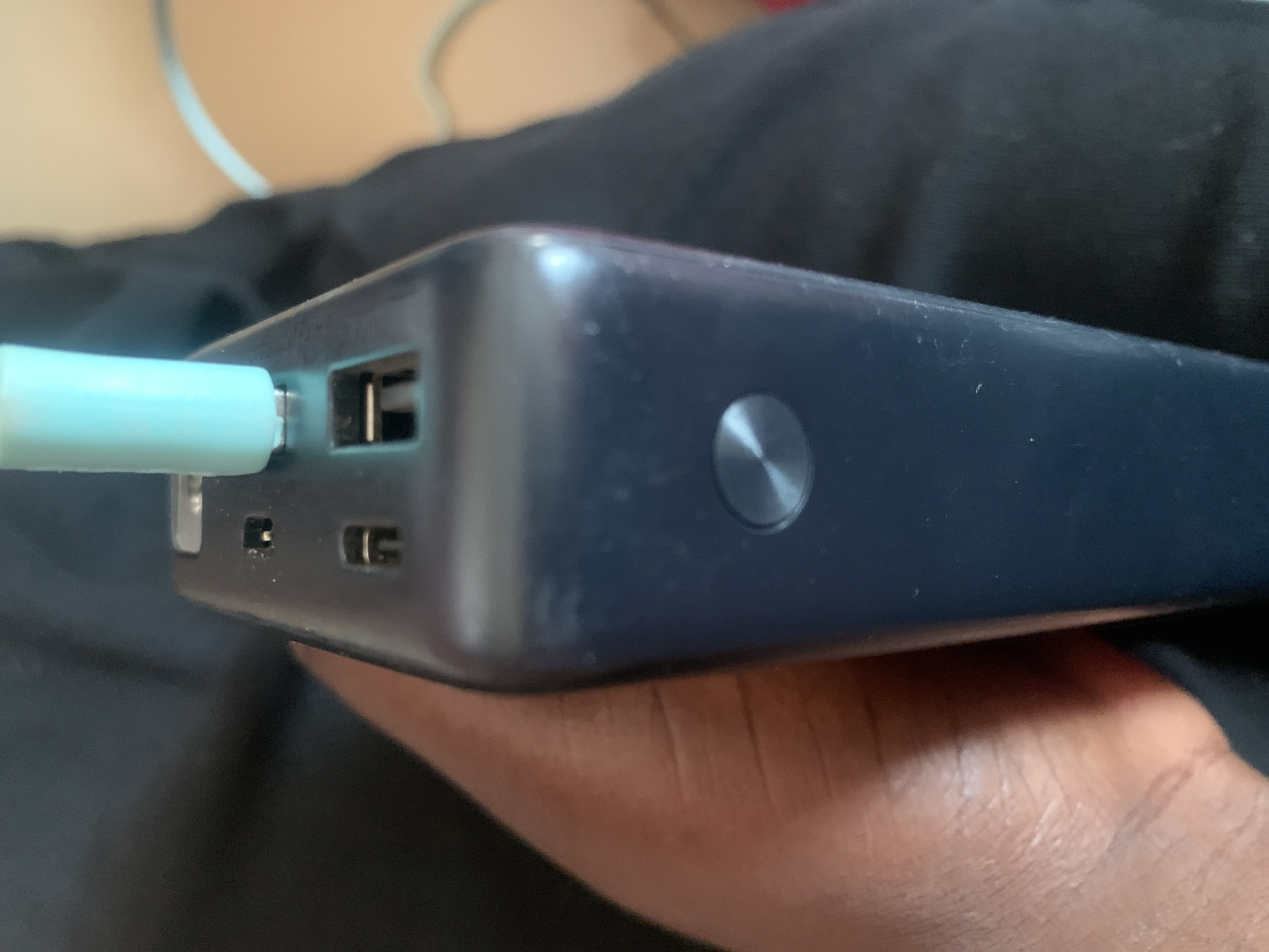 A close-up shot focuses on a dark blue power bank held in a person's hand. The power bank has a metallic circular button on its surface and several ports on its top. A light blue USB-C cable is plugged into one of these ports. The background is out of focus, showing dark fabric and a lighter-colored surface with a curved line. The setting appears to be indoors. No specific location in Jauro Yinu, Nigeria, can be determined from the image.
