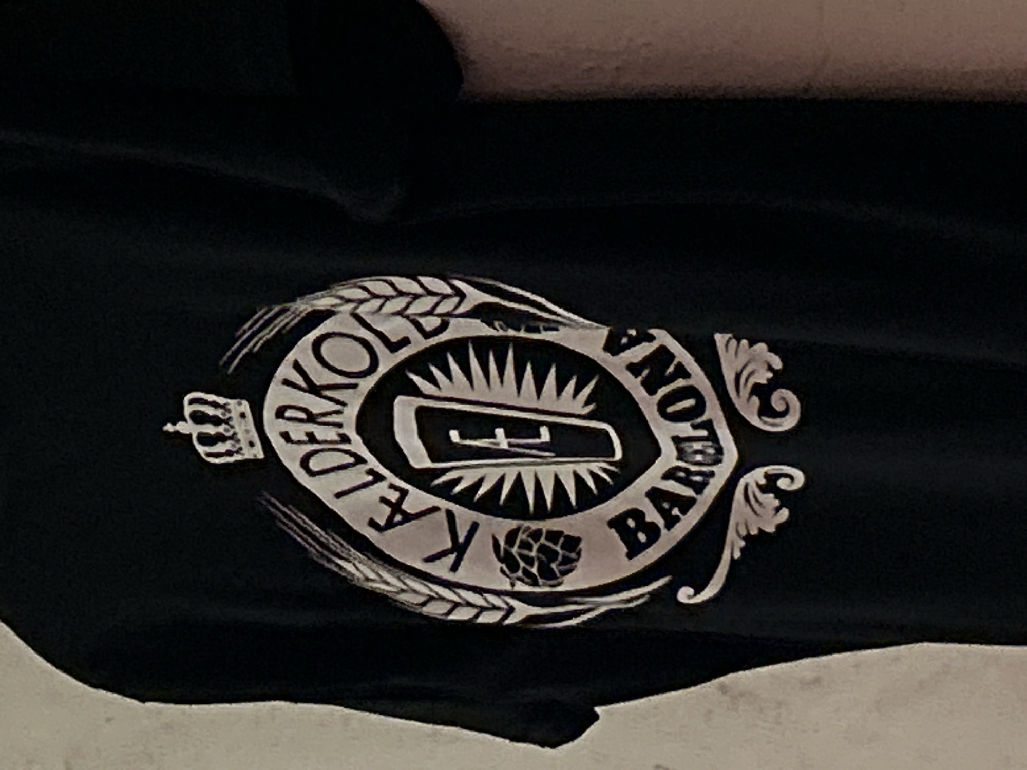 The image is a close-up of a black t-shirt with a white graphic design. The graphic is circular and features a crown at the top. Inside the circle, there are images of wheat stalks and a hop cone, with the words "KALDERKOL" written in an arc above a stylized beer mug. The bottom of the circle has the word "BARCELONA" written in an arc, with ornate scrollwork below it. The t-shirt is likely being worn by a person, but only the t-shirt itself is clearly visible. The background is a plain, textured wall, and a shadow is cast on the right side of the image, suggesting artificial lighting. There is no discernible information about the specific location within the city of Barcelona, Spain, as the image focuses solely on the t-shirt's design.