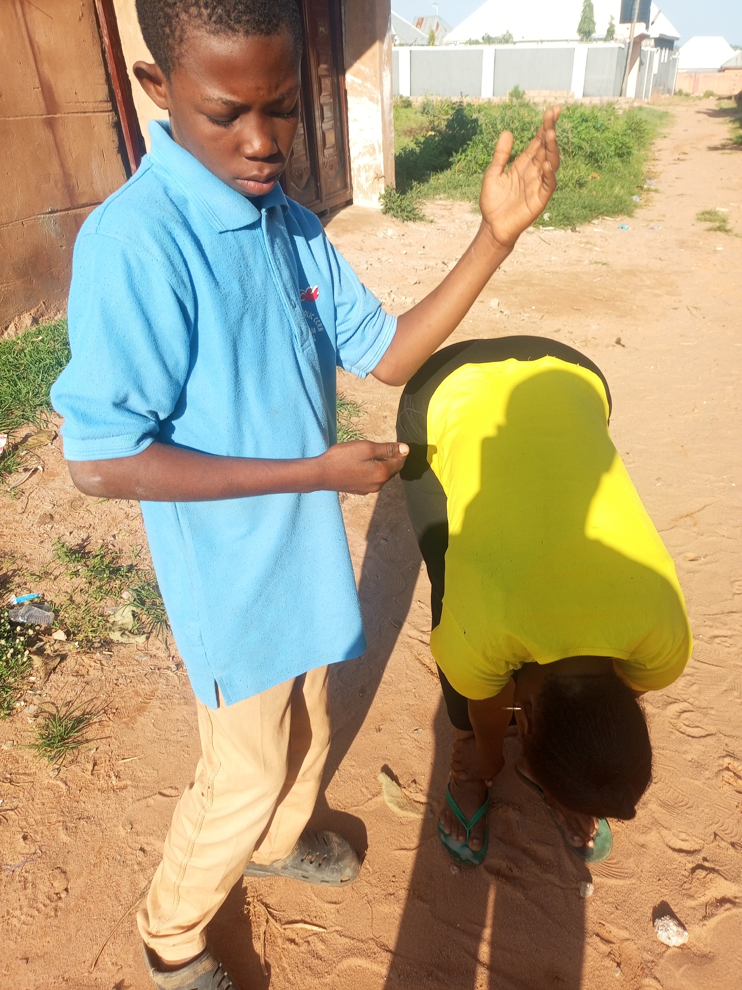 In the city of Tempure, Nigeria, two individuals are outdoors in what appears to be a dusty, unpaved area. The foreground is dominated by sandy ground, with patches of dry grass and small pieces of debris scattered about. To the left, a building with a brown, weathered facade is visible. In the background, low green vegetation lines a pathway, and beyond that, modern-looking buildings with gray walls and white roofs are discernible under a bright, clear sky.
A young boy, appearing to be of African descent, stands in the left-center of the frame, facing slightly to the right. He wears a bright blue polo shirt with a small emblem on the left chest and tan-colored trousers. His right arm is raised, palm open, in a gesture that could be interpreted as a greeting, a plea, or a signal. His gaze is directed downwards.
To the right of the boy, another individual, also of African descent, is bent over. This person wears a vibrant yellow t-shirt and black trousers. Their head is bowed, and they are wearing green flip-flops. The posture suggests they are either picking something up from the ground, attending to something on the ground, or perhaps in a pose of supplication or prayer. The interaction between the two is unclear, but their proximity and the boy's gesture suggest a connection. The lighting indicates it is daytime, casting distinct shadows on the sandy ground.
