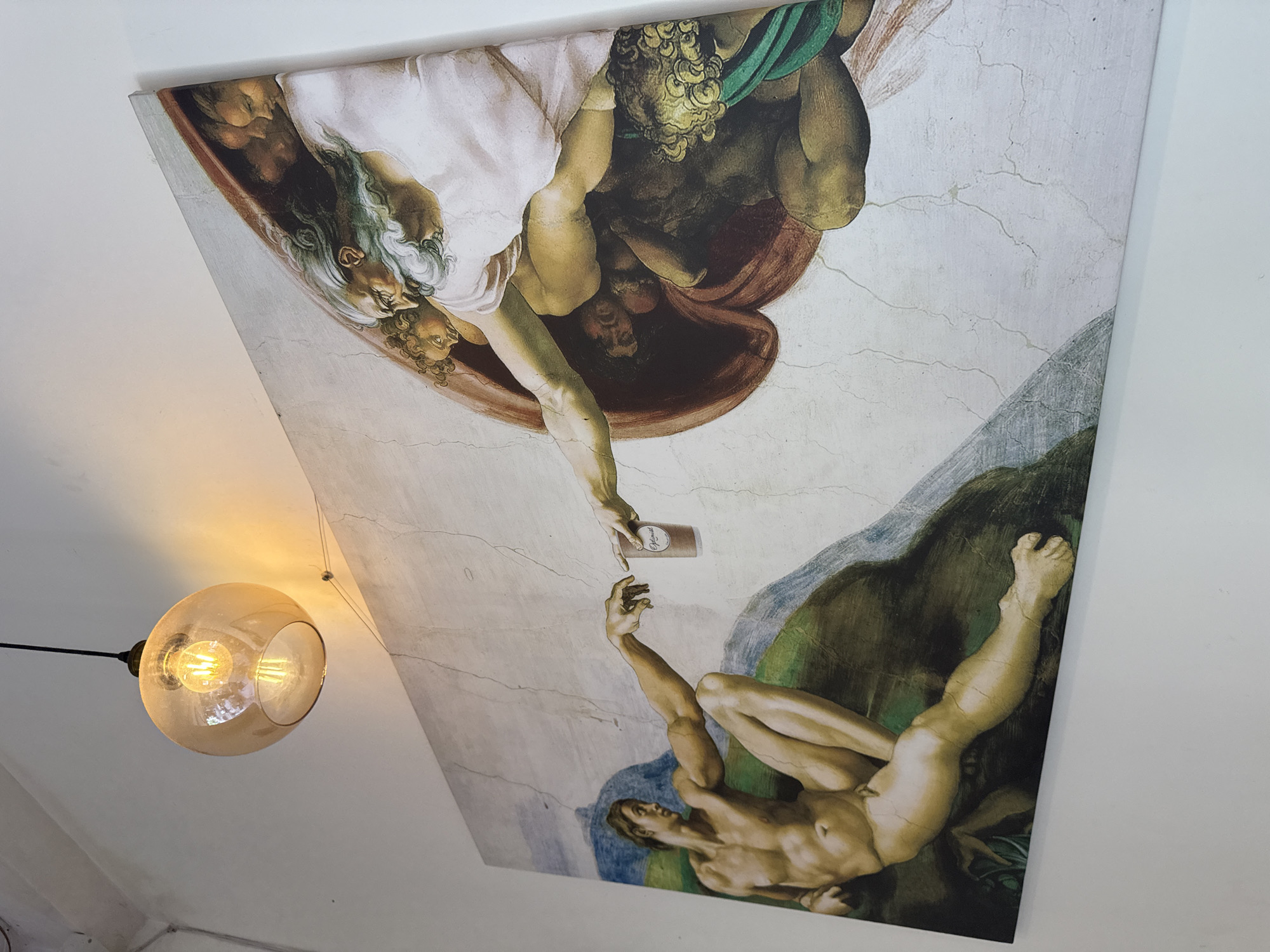The image displays a large canvas reproduction of Michelangelo's "The Creation of Adam" artwork, mounted on a white wall. The reproduction is altered; instead of Adam's finger touching God's finger, a brown coffee cup with the word "Optimist" in white lettering is held between their outstretched fingers. The cup appears to be a takeaway coffee cup. A warm-toned glass pendant light fixture hangs from the ceiling above the artwork, casting a soft glow on the scene. The setting is indoors, likely in a commercial establishment given the coffee cup modification. The overall impression is a humorous and modern twist on a classical masterpiece, possibly found in a cafe or restaurant. No specific location in Nha Trang Ward, Vietnam, is identifiable from the visual information provided.