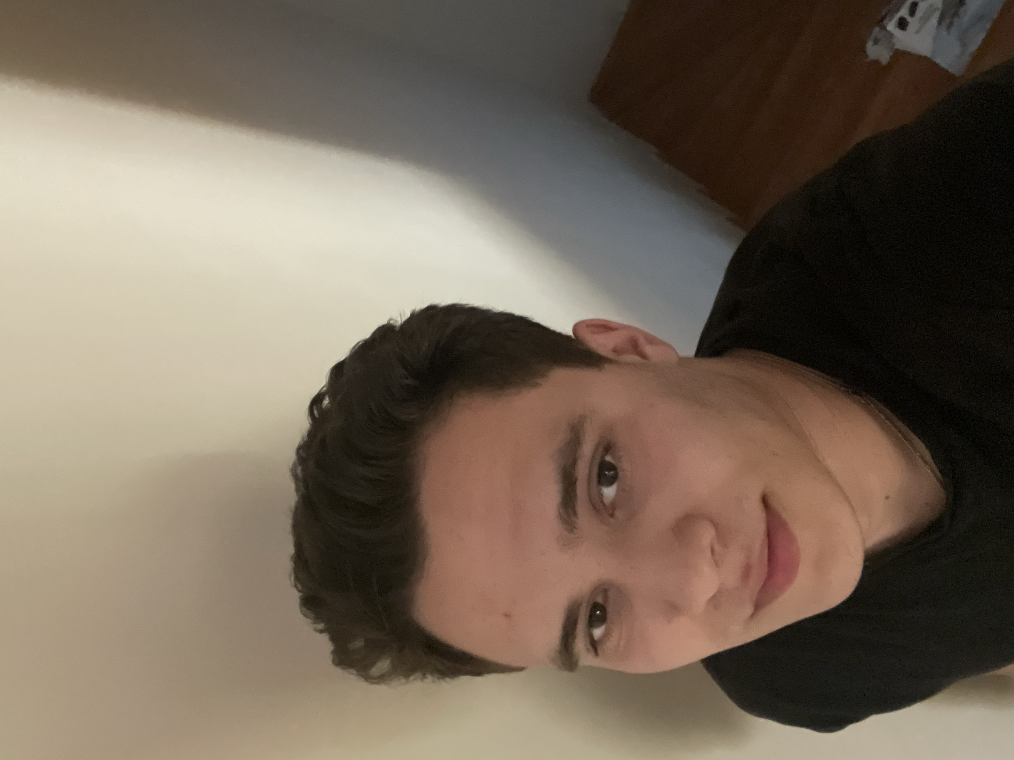 A young man with short, dark hair is looking at the camera. He is wearing a black shirt and appears to be indoors. The background consists of a light-colored wall and a wooden surface. The photo is tilted. There is no information suggesting that this photo was taken in Varna, Bulgaria.