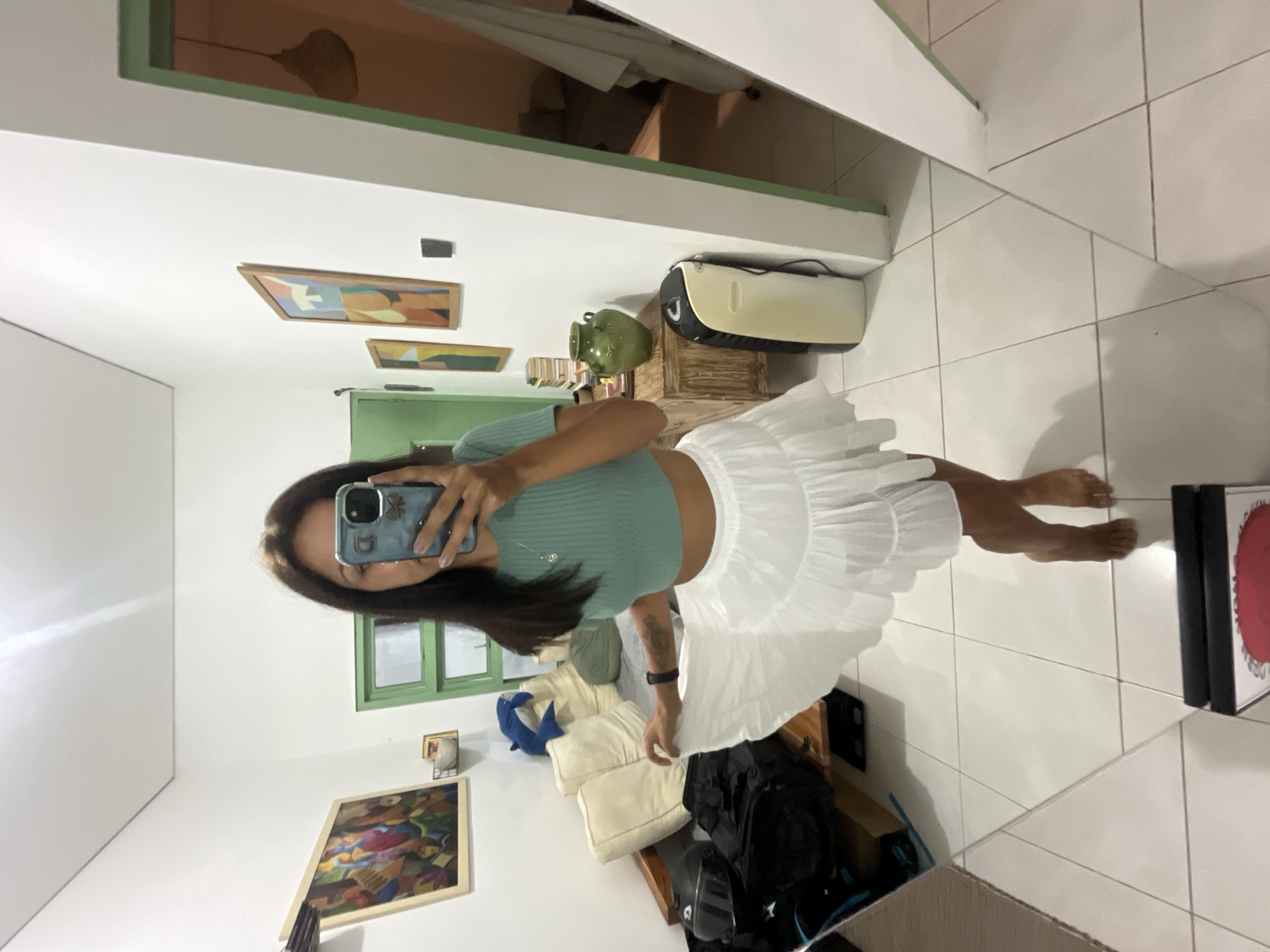 The image displays a mirror selfie taken by a person in an indoor setting. The subject, likely female, is captured from the waist up, holding a smartphone to take the picture. She is wearing a light green ribbed crop top and a white, ruffled mini skirt. A necklace with a charm is visible around her neck, and she has a watch or fitness tracker on her left wrist. Her dark hair falls over her shoulders, and her face is partially visible in the phone's reflection. The smartphone case is light blue with a floral or butterfly decal.
The background, reflected in the mirror, reveals a room with white walls and a white tiled floor. A distinctive green-painted window frame is visible. Several framed artworks adorn the walls, featuring abstract, geometric designs with varied colors including orange, blue, yellow, and green. Decorative elements include a green ceramic pot or vase, a cream-colored appliance (possibly a speaker) with black accents, and a blue stuffed animal (resembling a dolphin or shark) placed on what appears to be a wooden surface with white pillows. Other items in the reflection include black bags, possibly a helmet, and a rectangular object with a red circular design on the floor. A ceiling fan is also reflected overhead. The person is barefoot, and their feet are visible on the tiled floor. The scene appears to be well-lit, suggesting either daytime or good indoor lighting. The setting, located in Kuta Selatan, Indonesia, suggests a residential room or hotel space. No visible text is clearly discernible.