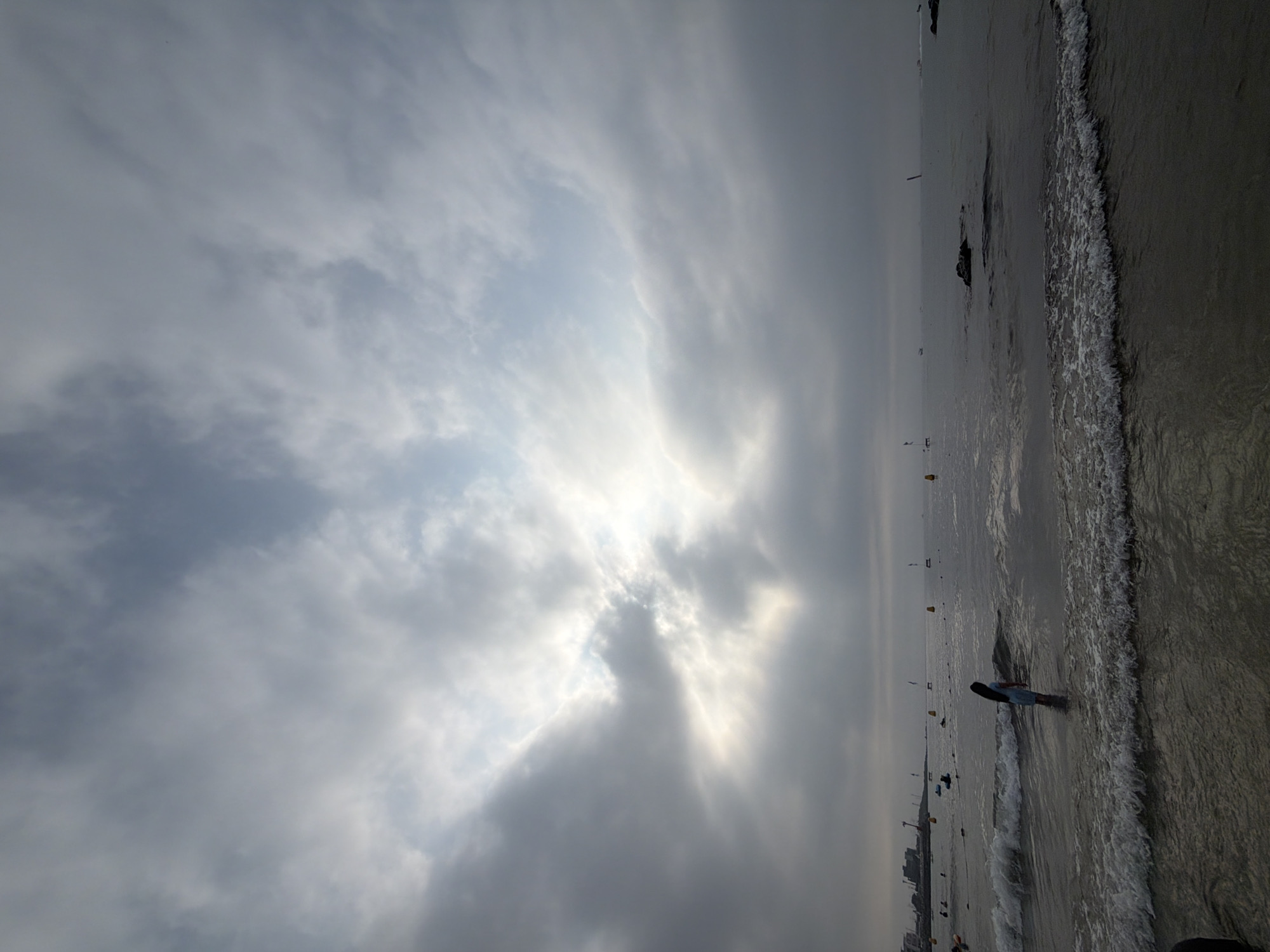 An overcast day at a beach in Jeju, South Korea. A lone individual, likely a young girl with long dark hair, stands in the shallow water, facing away from the camera. The sea is moderately active with waves breaking near the shore. In the distance, several buoys are visible in the water, and a hazy cityscape with buildings and construction cranes can be seen along the coastline. The sky is filled with heavy clouds, with a patch of brighter light breaking through near the horizon.
