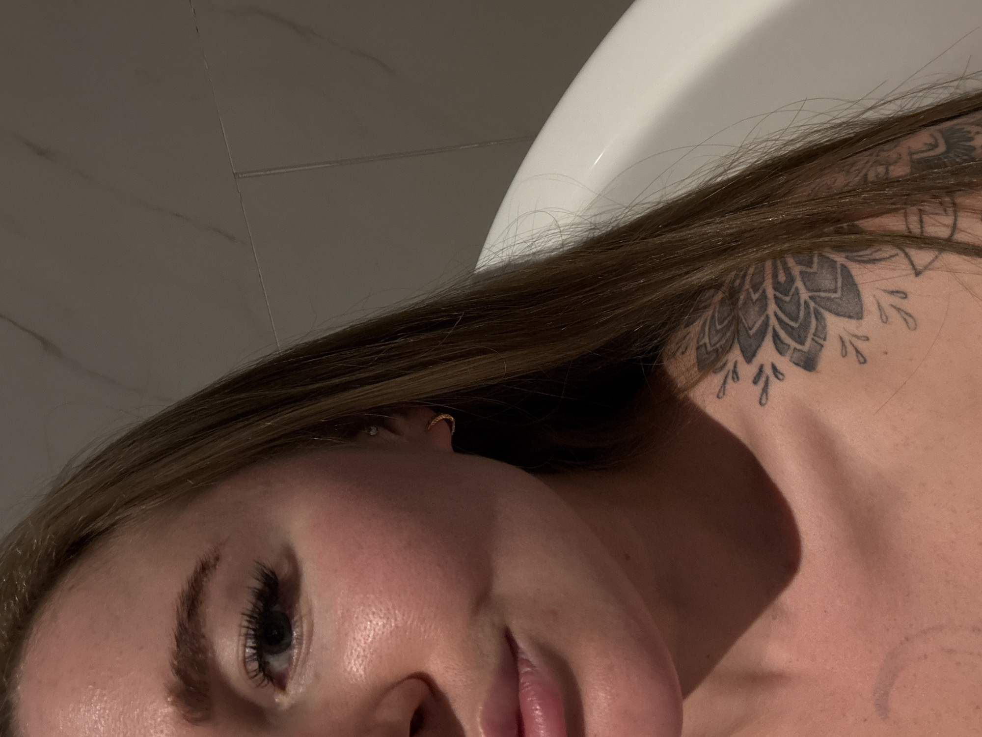 A close-up, angled photograph depicts a woman's face, neck, and upper chest. She has light skin, brown hair, and a tattoo of a geometric floral design on her shoulder. A gold earring is visible in her ear. Part of a white, curved object, possibly a bathtub, is visible in the background. The background also includes a textured, light-colored wall. The setting is a bathroom, but there is no indication of Zhangjiajie, China.