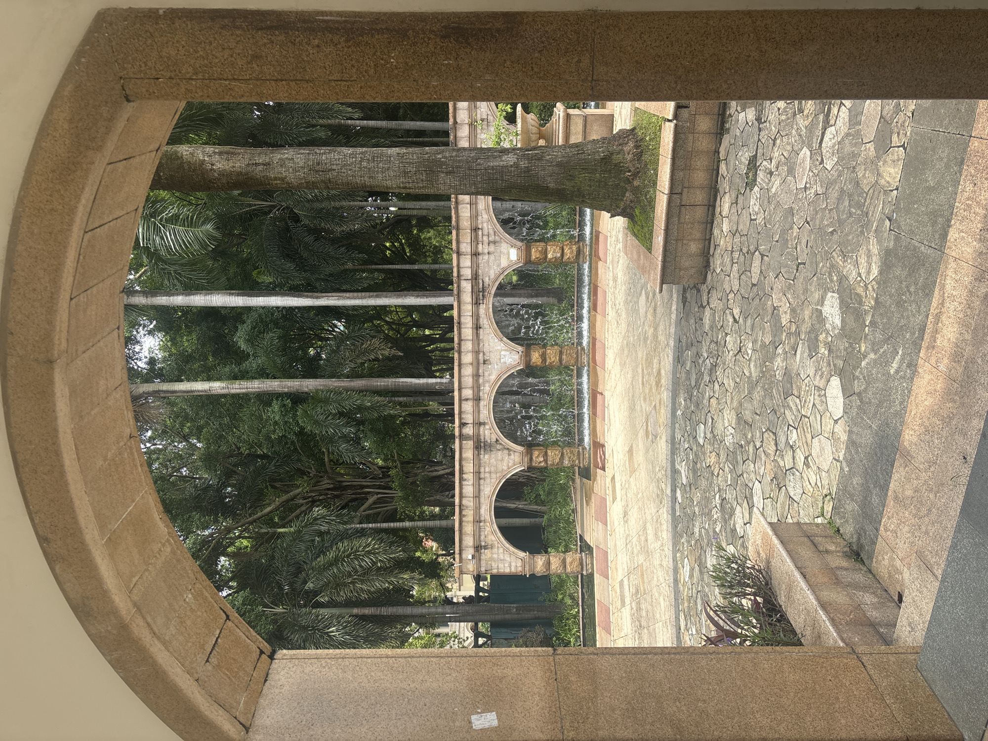 The image shows an arched entryway looking out onto a courtyard with a fountain and palm trees. The entryway is made of tan stone blocks. The courtyard is paved with irregular tan and gray stones. In the background, there is a wall with multiple arches where water cascades down. Beyond the fountain wall, tall palm trees and lush green foliage are visible. The overall impression is one of a serene, possibly tropical, outdoor space. The scene appears to be located in Nanhai District, China. No people are visible in the image.