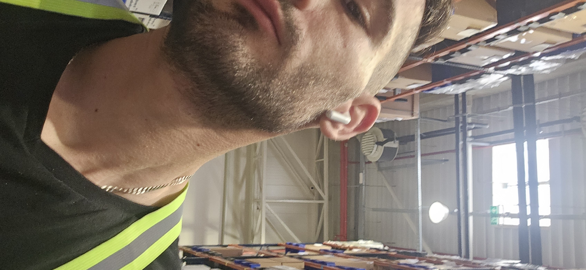 A close-up, slightly tilted, indoor shot depicts a man in a black shirt with a green and gray reflective stripe, wearing a silver chain necklace and a white wireless earbud in his right ear. He has a beard and is gazing out of frame. The background is a warehouse interior. A large fan is visible on the wall, along with red pipes and white walls with metal supports. Light streams in from a distant window. Overhead are metal warehouse racks, some with boxes stacked on them. Pallets, some with boxes, are scattered on the warehouse floor. The image lacks any contextual information to determine if it was taken in Plovdiv, Bulgaria.