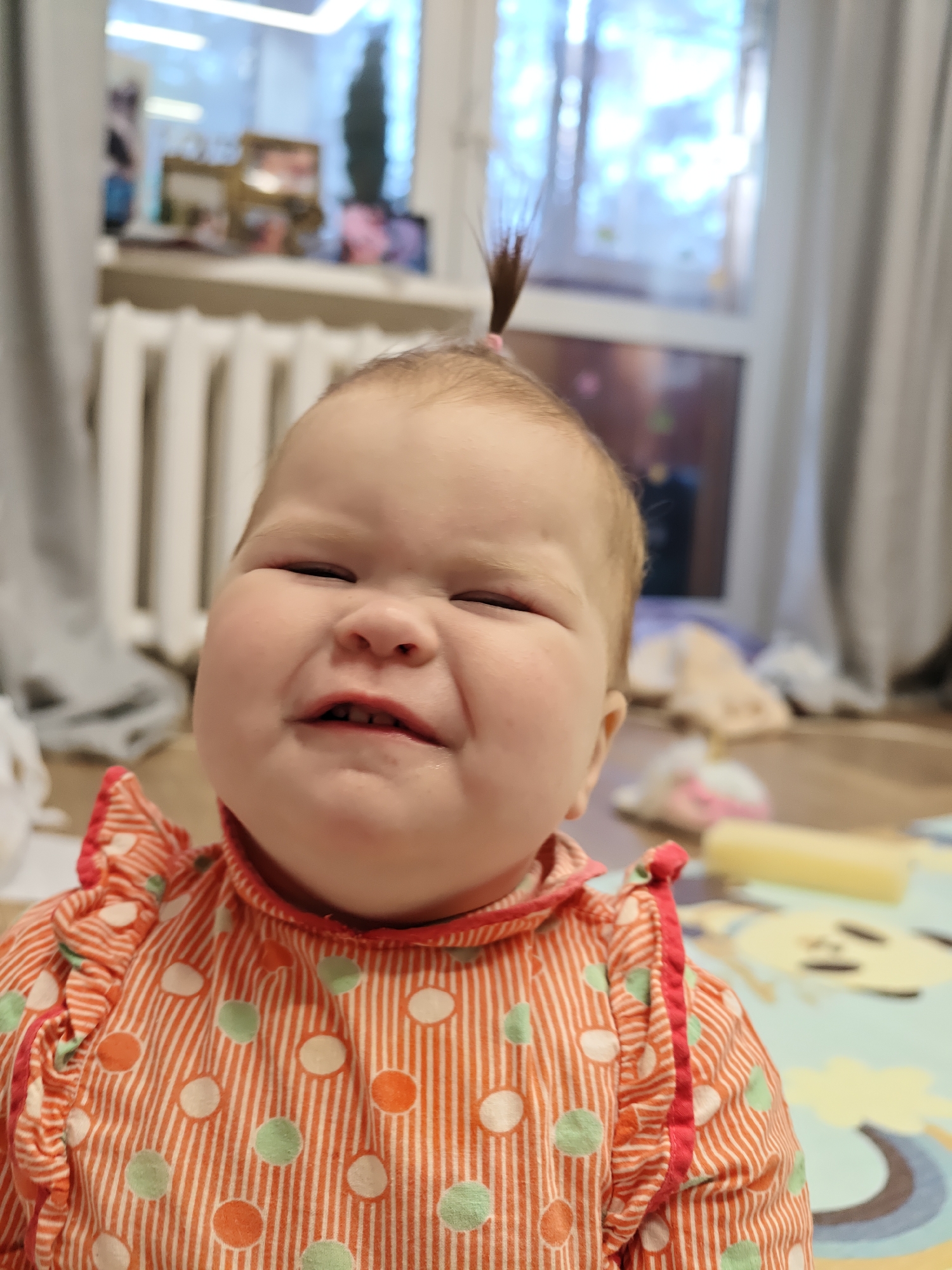 A close-up, slightly blurred shot depicts a baby girl making a face. The baby has a small tuft of hair tied on top of her head. She is wearing an orange and white striped shirt with green and orange polka dots and red trim with ruffles around the collar and sleeves. The background shows a radiator, a window, and what appears to be a play mat on the floor. The location is potentially a residence in the city of Chaikovsky, Russia.