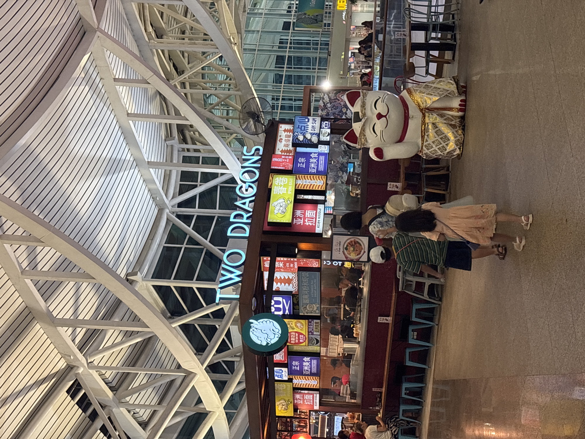 The photograph captures the "Two Dragons" restaurant, with signage in both English and Chinese, within an airport terminal. The building's architectural structure features a white, angular framework that supports a curved roof. The restaurant's exterior showcases a brightly lit menu board and a large, seated, waving Maneki Neko figure. Customers are visible at the restaurant's counter, and people are walking past. Based on the presence of the "Kuta, Indonesia" tag and the architecture, it can be inferred that this image was taken at the Bali Ngurah Rai International Airport in Kuta, Indonesia.