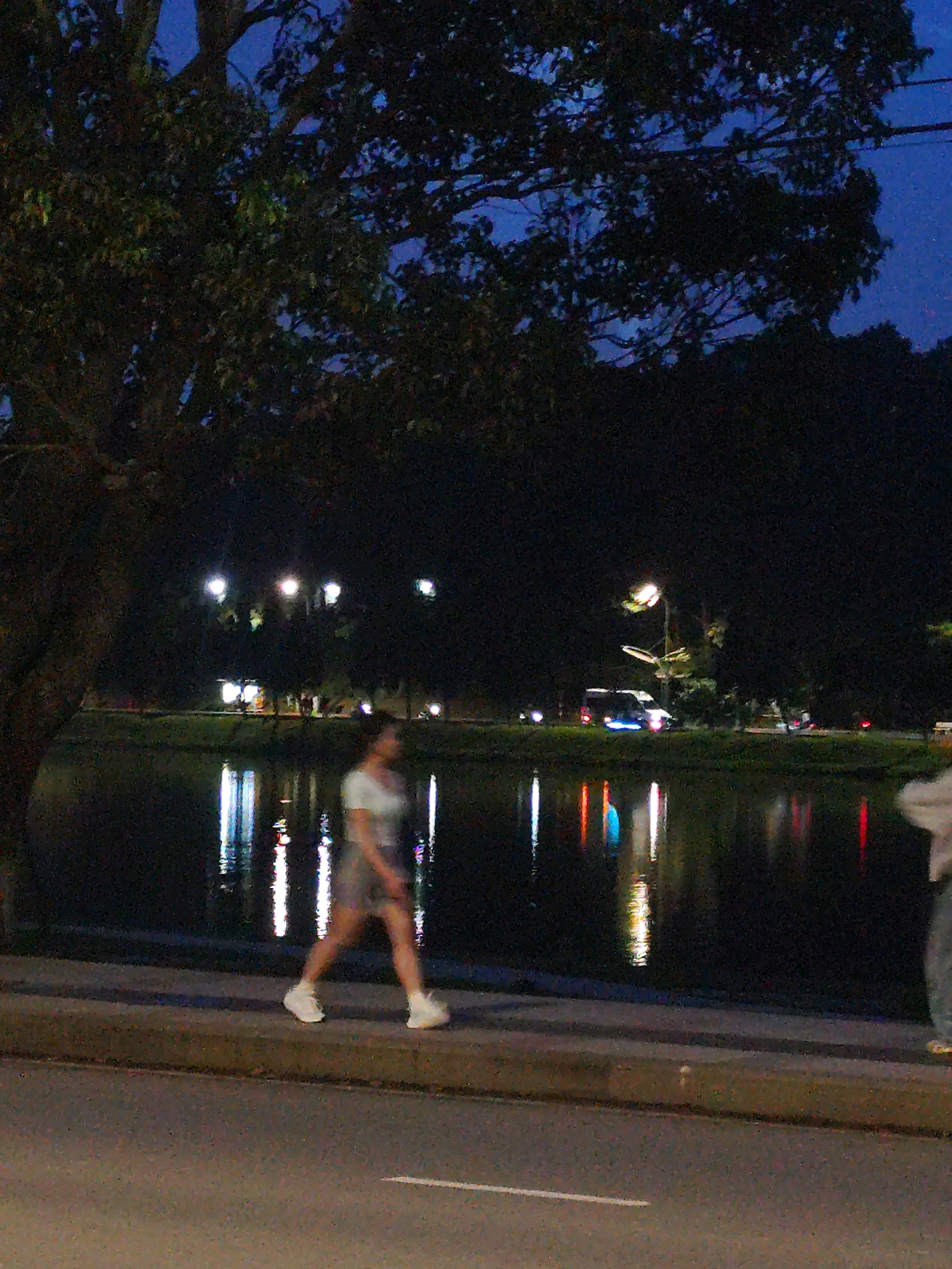 At dusk, a solitary young woman walks along the paved pathway bordering Xuan Huong Lake in Da Lat, Vietnam. She wears a light-colored t-shirt and shorts, with white sneakers. The dark water of the lake reflects the lights from street lamps and passing vehicles. The background reveals the silhouette of trees and hills. The overall atmosphere is quiet and serene, indicative of an evening stroll in a park-like setting.