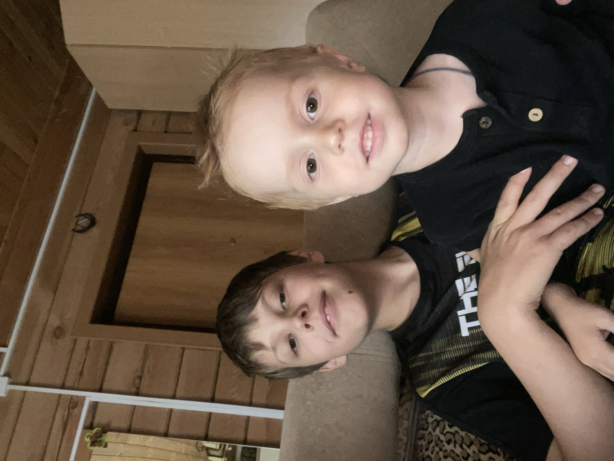 The image, taken in Chaikovsky, Russia, features two young boys posing for a photo indoors, presented rotated 90 degrees clockwise. Both boys are seated or reclining on a light brown, upholstered couch or armchair, looking towards the camera with gentle smiles.
The younger boy, with blonde hair and fair skin, is positioned in the foreground (left side when viewed upright). He is wearing a black collared shirt with two light-colored buttons. A thin, dark cord or bracelet is visible on his neck/shoulder area.
The older boy, with brown hair, is situated behind and to the right of the younger boy. He is dressed in a black t-shirt featuring a yellow and black pattern, possibly camouflage or geometric, and some white text. The letters "T" and "H" are partially visible on his shirt. His hands are resting in front of him. A patterned rug or blanket, possibly leopard print, is visible beneath him on the couch.
The background consists of wooden paneled walls, suggesting a rustic or cabin-like interior. A wooden door frame leading into a dark opening is visible in the middle background. A dark, horseshoe-shaped object hangs on the wooden wall, and a white horizontal bar or rail runs across the lower part of the wall. The lighting suggests it is daytime, and the overall mood is calm and friendly, conveyed by the boys' expressions.