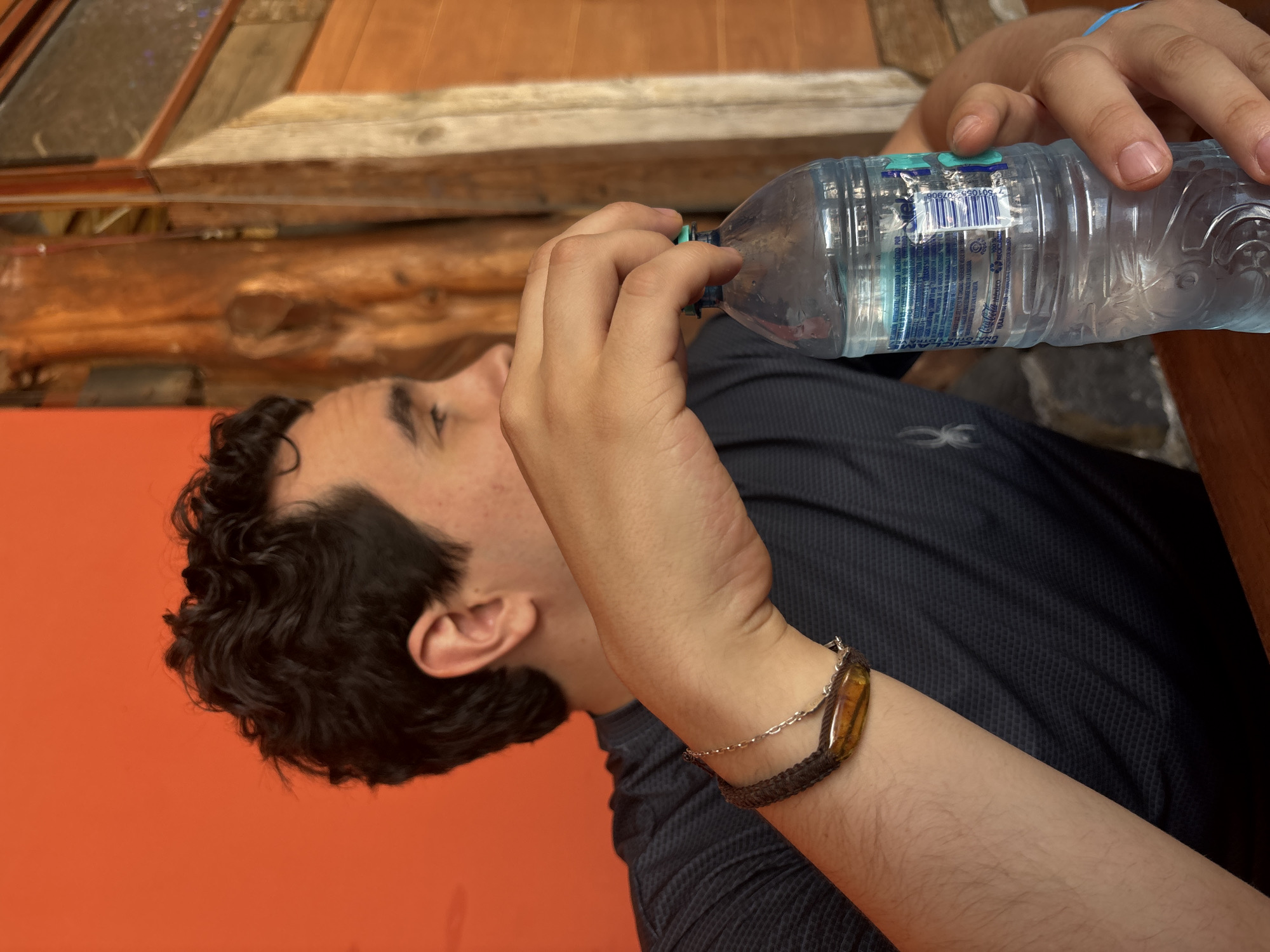 A light-skinned man is drinking from a clear plastic water bottle. He has dark curly hair, with a wrist adorned with a woven bracelet with a tan stone. He is wearing a dark blue shirt. The background features an orange wall and a wooden door frame with textured brown wood. This is taken at the location San Juan Arcos Ojo de Agua in Mexico.