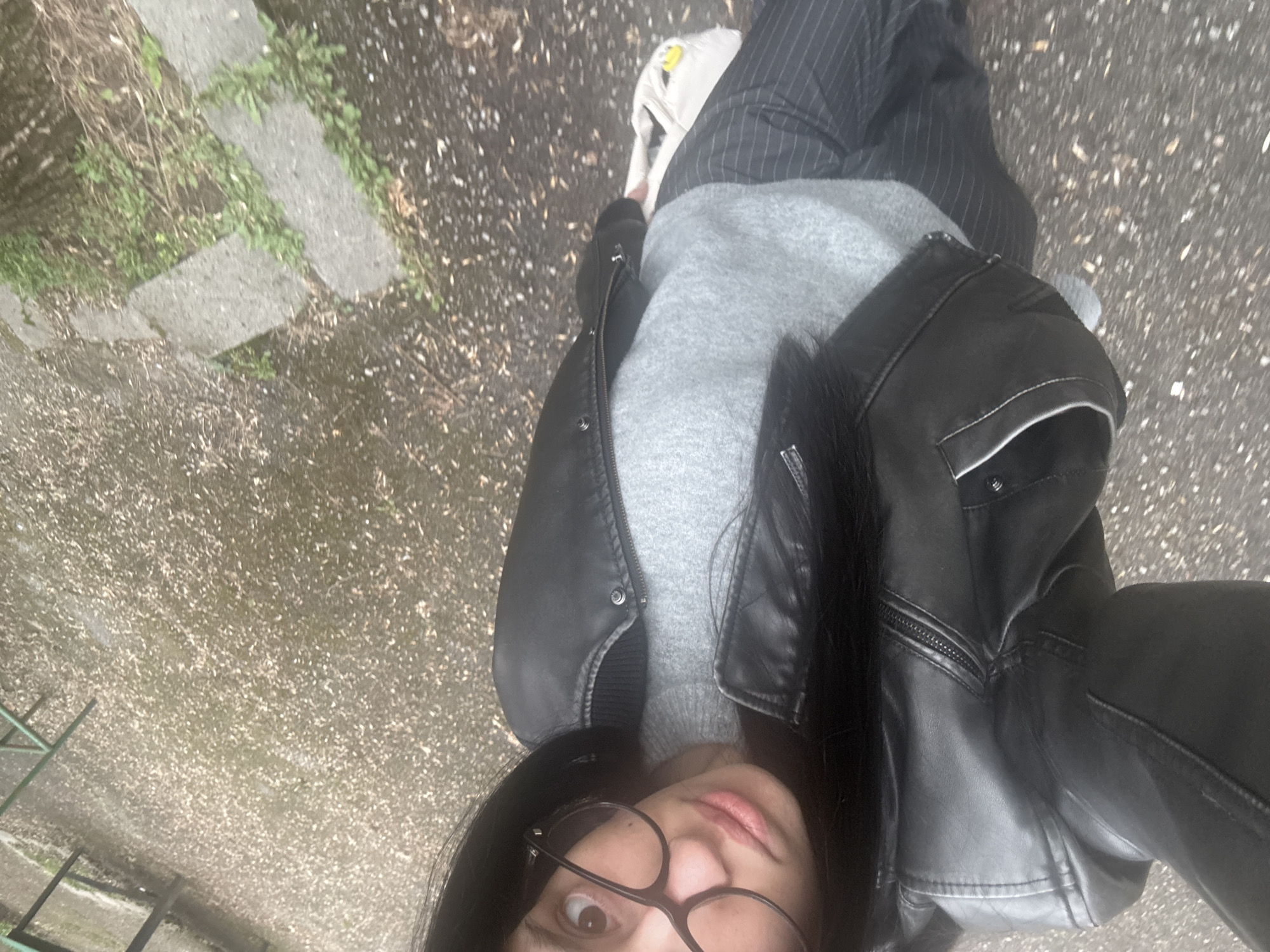 A person with dark hair and glasses is taking a selfie outdoors. The person is wearing a black leather jacket over a grey sweater and striped pants. A white bag is visible near their legs. The background shows a paved surface with some greenery and stones. Parts of what appears to be a playground structure are visible in the lower left corner. Given the style of architecture, paving, and playground equipment, the location might be in Sofia, Bulgaria.