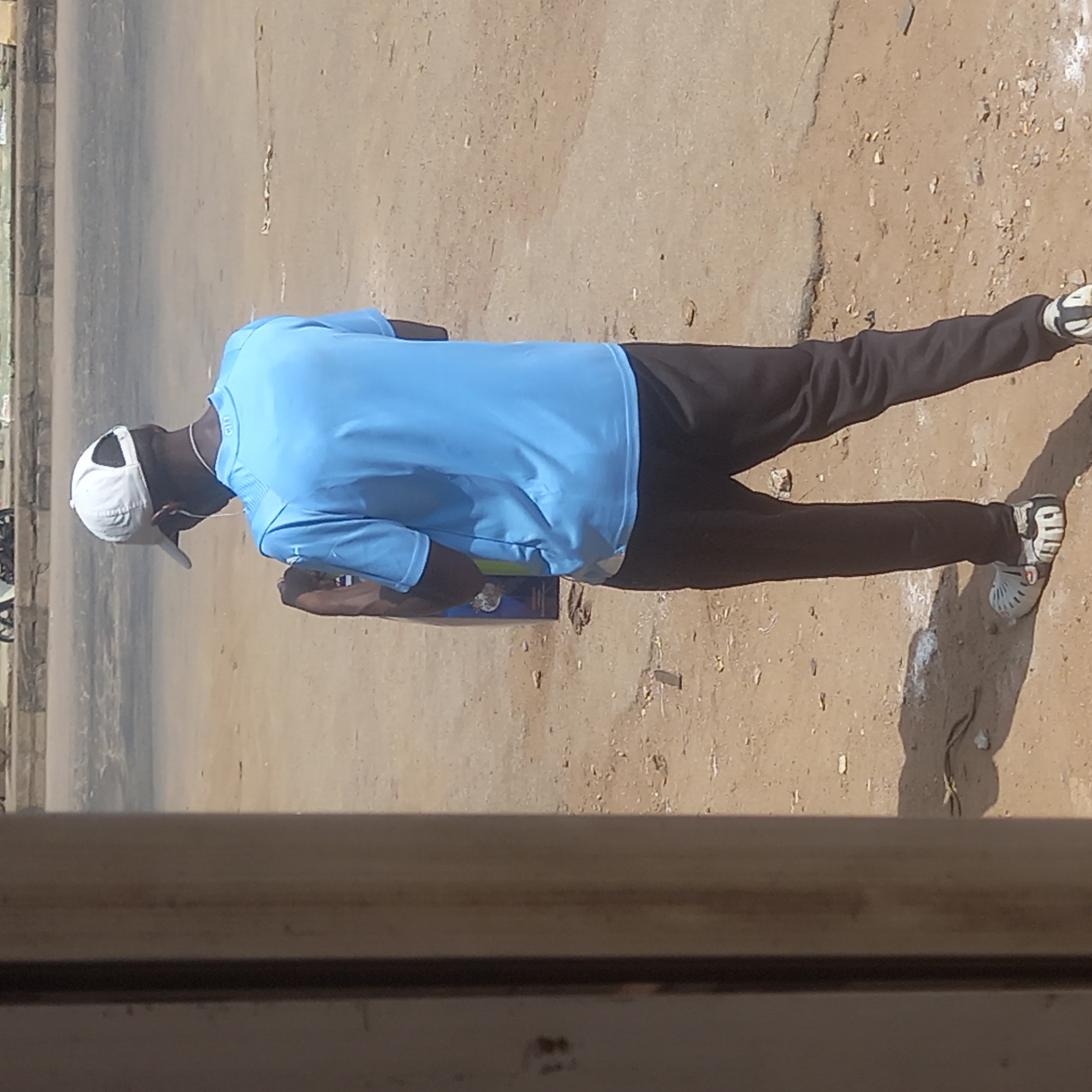 A man wearing a light blue shirt, dark trousers, white hat and sandals stands on a dirt road next to a paved road in Dadi Riba, Nigeria. He is holding a dark object in his hands. The scene is viewed through a window frame.