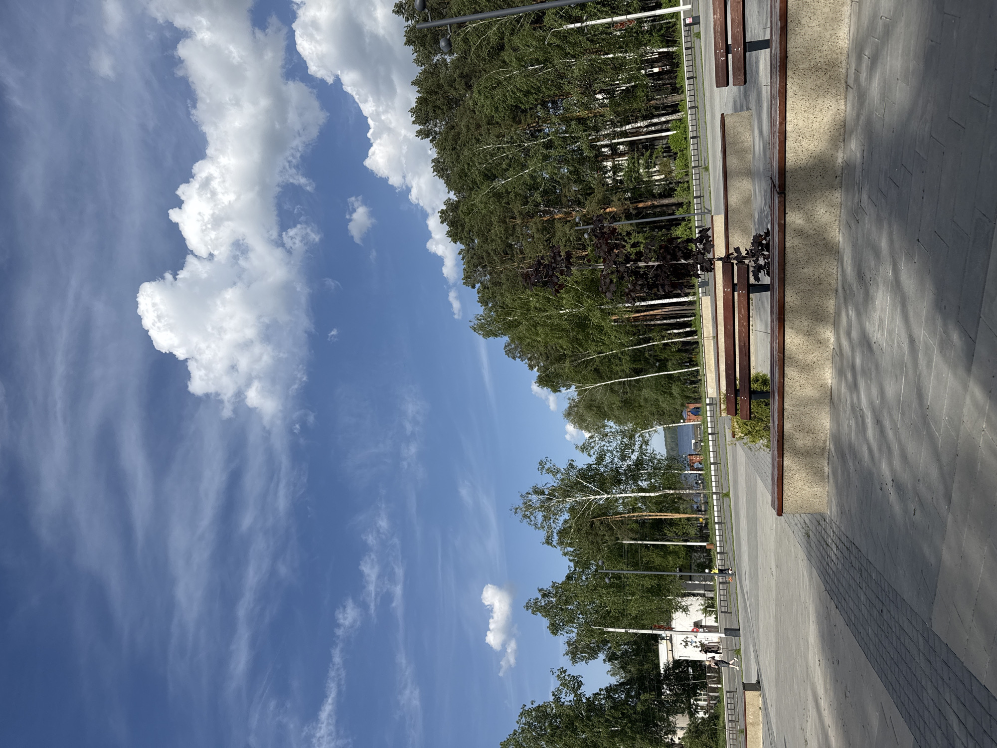 The image depicts a park scene in Chaikovsky, Russia. The sky is blue with scattered cumulus and cirrus clouds. Along a paved walkway, there are multiple benches with a combination of wood and stone elements. A line of trees runs parallel to the walkway, including birch trees. Lighting poles are positioned among the trees. Distant buildings and a body of water can be seen through the trees. The walkway appears to be empty of people in the immediate foreground, but there are indistinct figures of people and possibly vehicles farther in the distance. The lighting suggests it is daytime with clear weather.