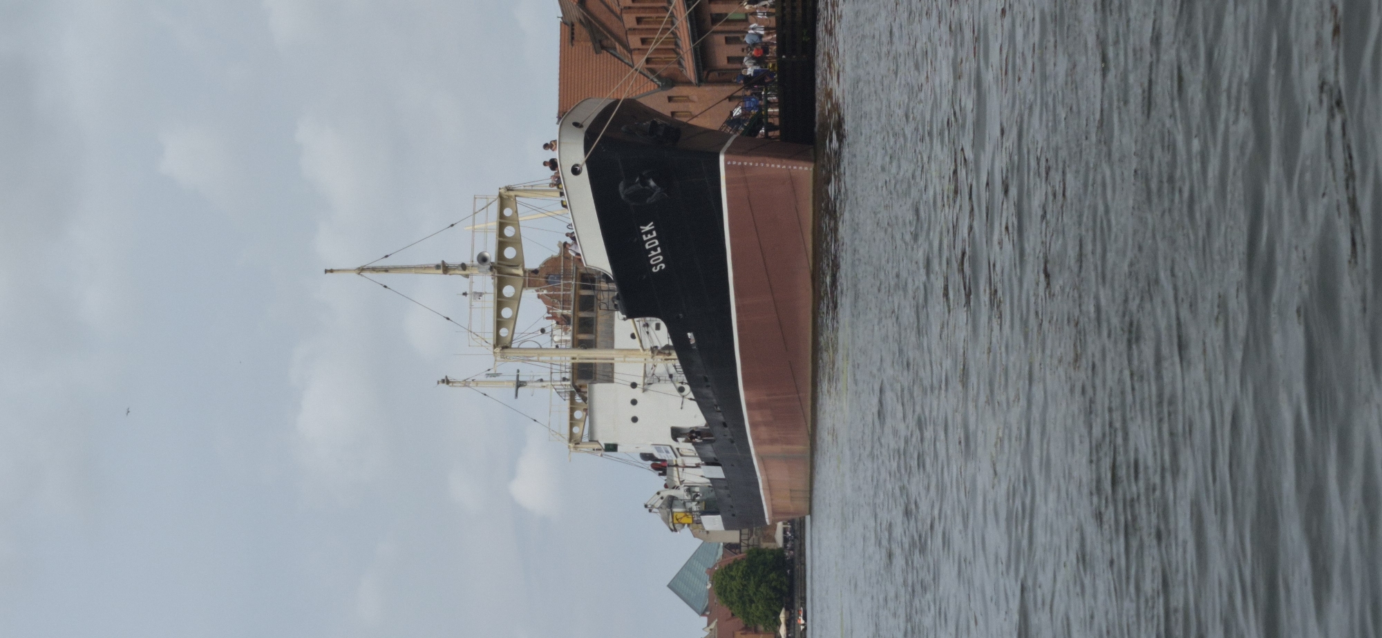 A motorship named "SOŁDEK" is docked in the Motława River in Gdańsk, Poland. The ship, painted black with a red hull, has a white superstructure with several decks and masts. A group of people are visible on the deck and along the pier. In the background, there are buildings with red tile roofs and a triangular green structure. The water is calm, with ripples indicating a slight breeze. The sky is overcast. The scene captures a moment in the port of Gdańsk, likely a tourist attraction given the presence of visitors on the ship and the well-maintained buildings along the riverfront.