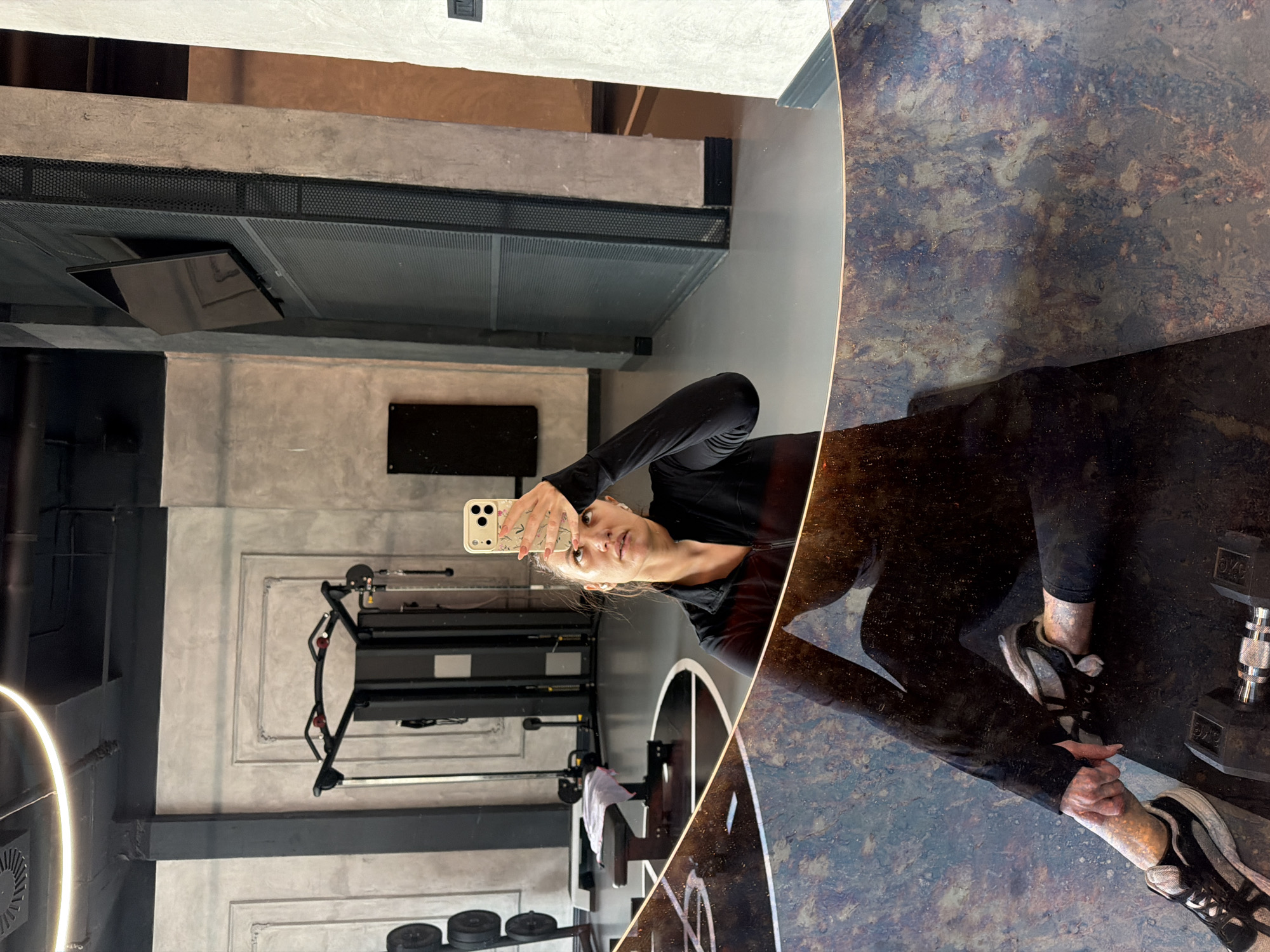 A woman in black athletic wear stands in a gym, reflected in a dark, possibly marble or stone, surface. She holds a smartphone up, presumably taking a selfie. In the background, gym equipment including a weight rack and dumbbells can be seen. The gym’s interior features a mix of gray concrete and black metal accents. The exact location is a gym in Belgrade, Serbia.