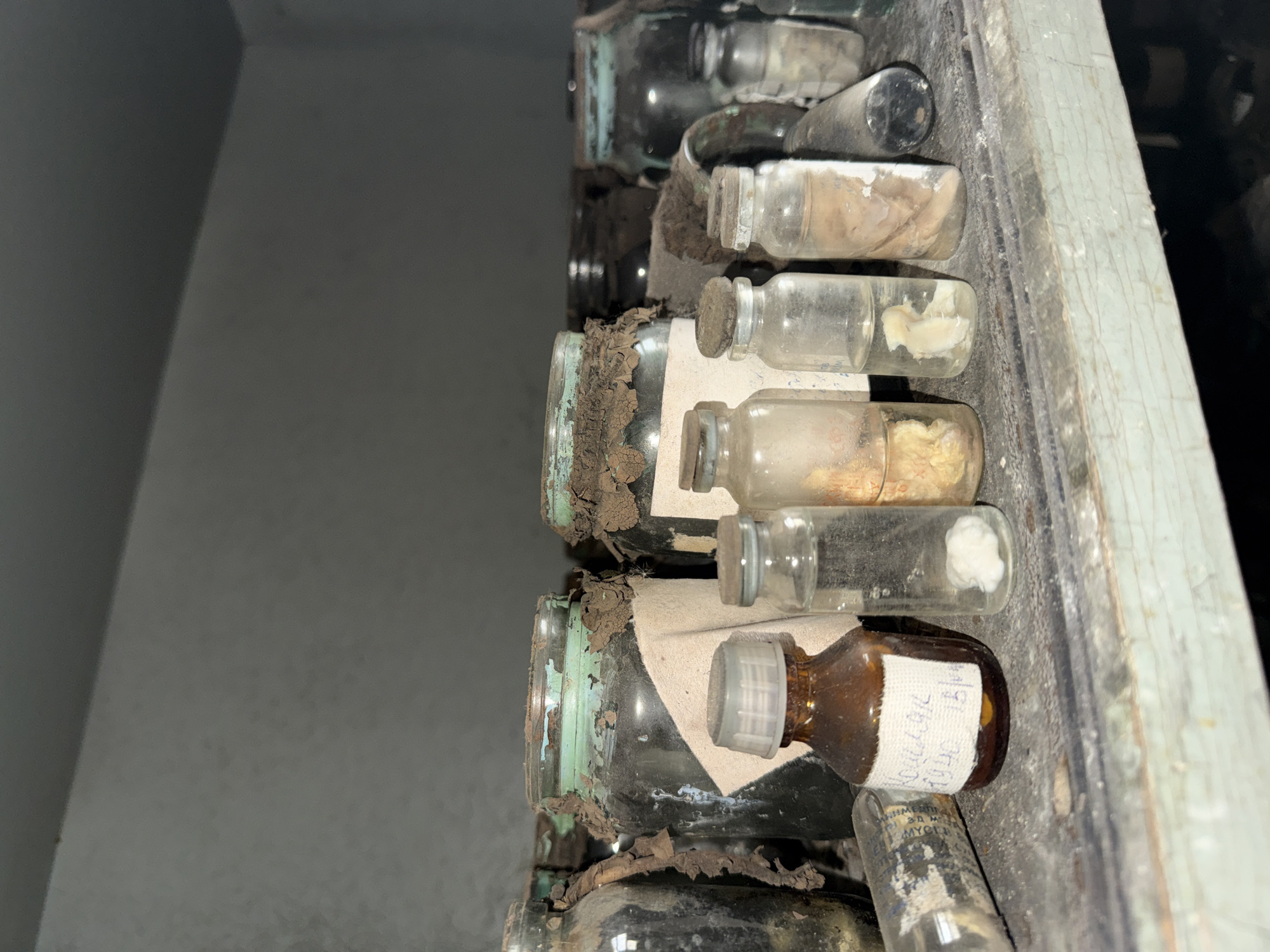 The image shows a collection of old jars and bottles filled with unidentified substances, arranged on a shelf. The containers appear to be of various sizes and shapes, with some having labels that are partially illegible. The substances inside the jars and bottles also vary in appearance, ranging from liquids to solids. The shelf and containers are covered in dust and grime, indicating that they have been left undisturbed for a long period of time. The environment is dimly lit and the overall scene suggests an abandoned or neglected storage area. Considering the location in Prypiat, Ukraine, it is likely that these items were left behind following the Chernobyl disaster, and the jars and bottles might contain specimens of biological or chemical substances. The condition of the containers suggests a lack of maintenance and deterioration over time due to exposure to the elements.