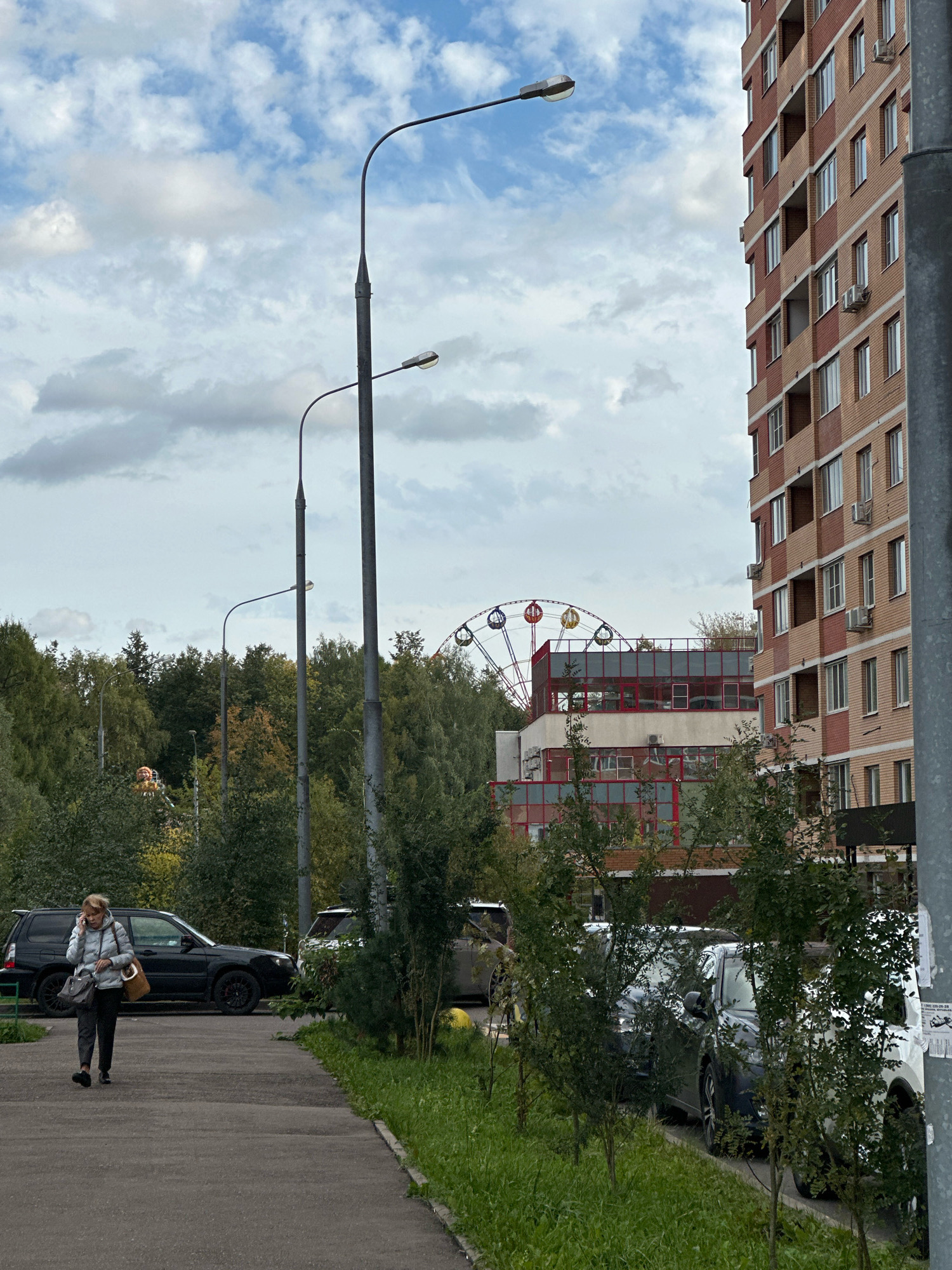 An outdoor, eye-level, full shot captures a residential street scene in Vidnoye, Russia, under a cloudy sky. A woman in a light-colored jacket and dark pants walks on a paved sidewalk to the left, talking on her mobile phone, carrying a tan shoulder bag and a dark crossbody bag. Adjacent to the sidewalk is a grassy area and a row of parked cars, including a black SUV. In the background, a multi-story apartment building with brick and light-colored siding stands to the right. Further back, a Ferris wheel and a building with a red-trimmed roof are visible behind a line of trees. Multiple streetlights are positioned throughout the scene.