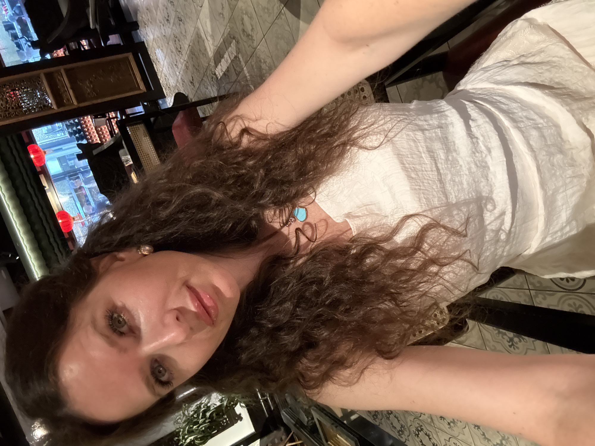 The image presents a female subject taking a selfie within an upscale Asian-themed restaurant in Dubai, United Arab Emirates. The subject has long, wavy dark brown hair, hazel eyes, and is wearing a white textured dress or top, a gold-toned stud earring in her visible right ear, and a necklace with a gold initial pendant and a turquoise stone. She maintains direct eye contact with the camera, exhibiting a slight smile. The background shows a dimly lit restaurant interior featuring dark wooden furniture, an ornate wooden screen with intricate carvings, and a geometrically patterned tiled floor. Through large windows or glass partitions, multiple glowing red Chinese lanterns are visible, behind which a blurred urban nighttime landscape, consistent with city buildings and lights, can be discerned. The specific architectural style, particularly the red lanterns and ornate woodwork, strongly suggests a high-end establishment such as Hutong in the Dubai International Financial Centre (DIFC).
