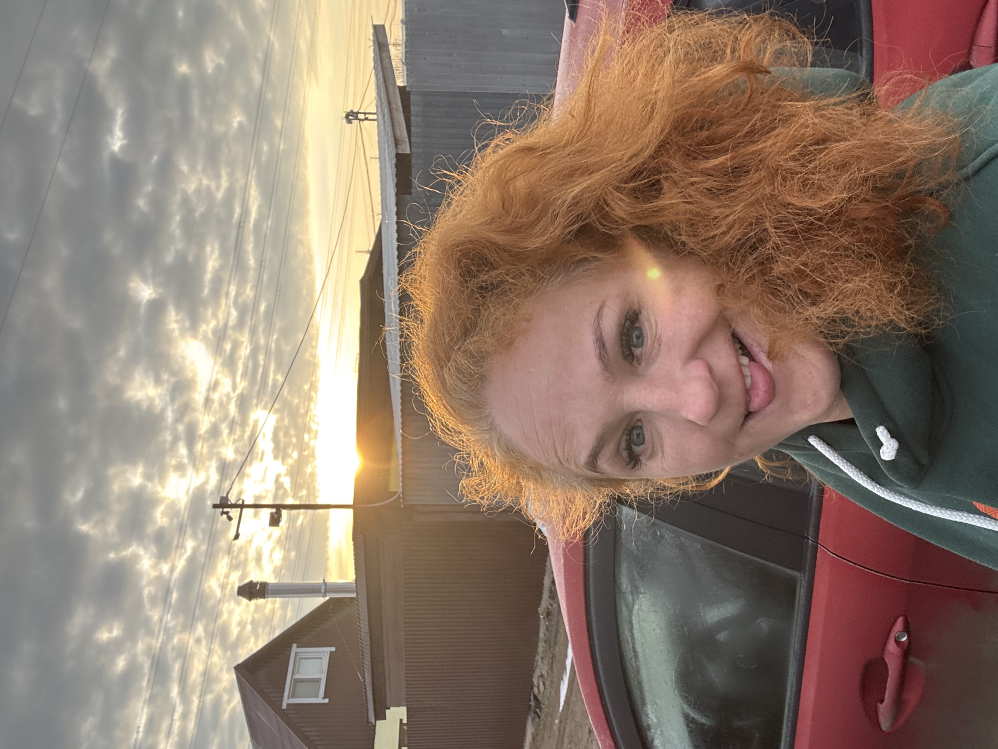The image is a selfie of a woman with red curly hair, light skin, and blue eyes. She's sticking her tongue out and smiling. She's wearing a green hoodie with a white drawstring. The photo appears to be taken outdoors near a red car. In the background is a house with a chimney, power lines, and a cloudy sky with the sun setting.
It is impossible to confirm this image was taken in Chaikovsky, Russia without additional information from the picture.