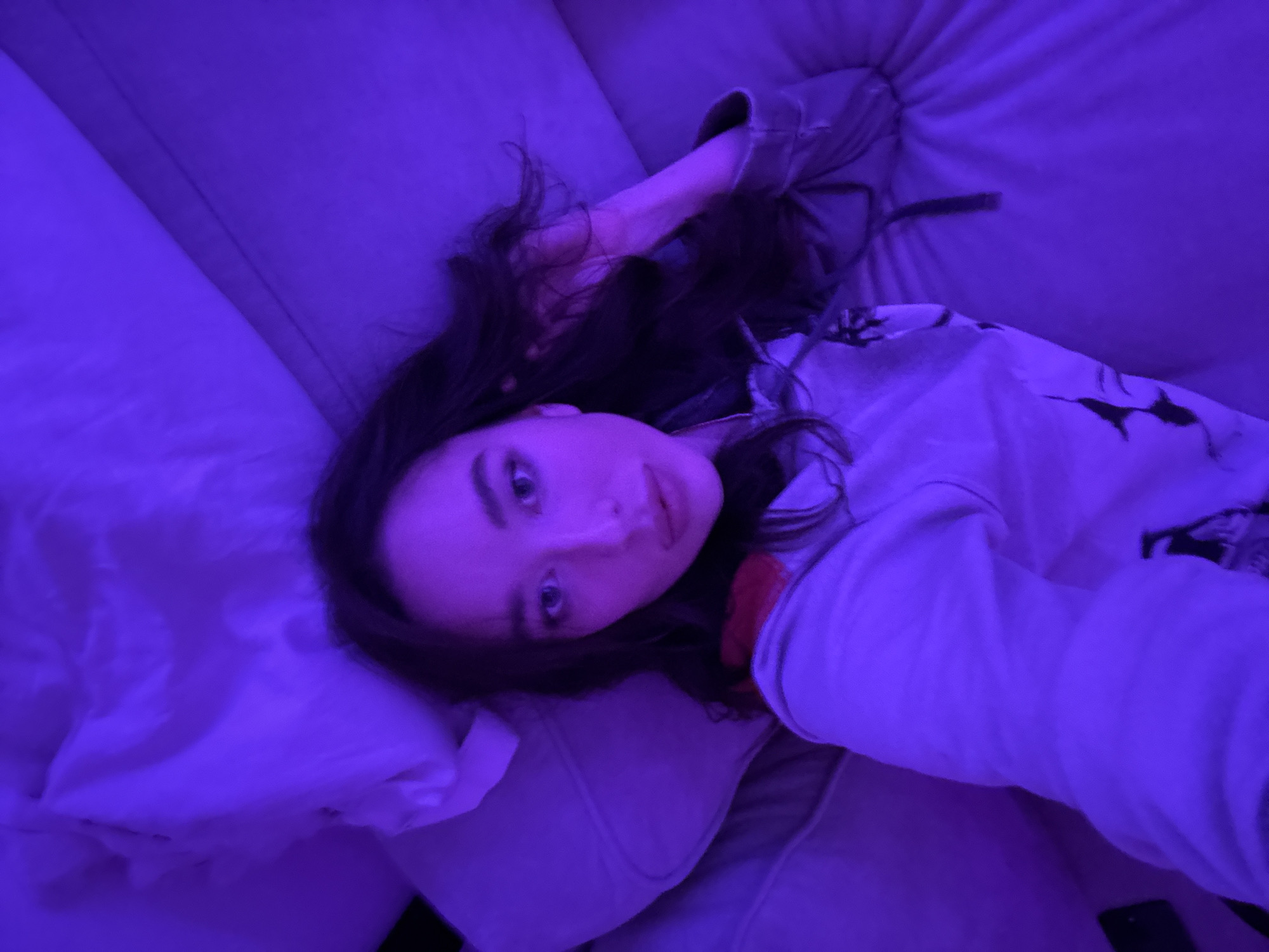 The image captures a young woman lying on a light-colored sofa or bed, illuminated predominantly by an artificial purple light. She is the central subject, positioned with her head slightly to the left and her body extending towards the right. Her long, dark hair is spread around her head and shoulders. She looks directly at the camera with a calm and serene expression; her eyes are light-colored, and her lips are slightly parted.
She is dressed in a light-colored, long-sleeved top, possibly a hoodie, which features subtle dark patterns visible on the fabric near her right shoulder and arm. Her head rests on a pillow, which blends with the surrounding light-colored upholstery of the furniture. The strong violet hue of the lighting envelops the entire scene, creating a distinct, moody, and intimate atmosphere, suggesting an indoor setting likely during the evening or night. There are no specific architectural or environmental cues within the image itself to indicate a particular location, although the provided context suggests Moscow, Russia. No visible text is present in the media. The activity appears to be a casual selfie or a moment of relaxation.